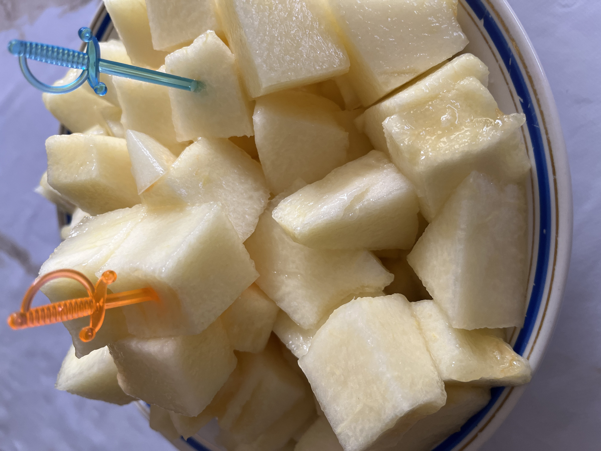 A close-up, eye-level shot shows a white ceramic bowl filled with cubed honeydew melon. The melon is cut into bite-sized pieces and appears juicy, with some pieces glistening. Two plastic sword-shaped picks are inserted into the melon; one is orange and the other is blue. The bowl has a decorative rim with blue and gold stripes. The background is blurred and appears to be a light-colored surface. The image depicts a prepared dish of fruit, likely a dessert or snack, presented in a bowl. The location in the city of Krasnyye Barrikady, Russia, is not discernible from the visual elements present in the image.