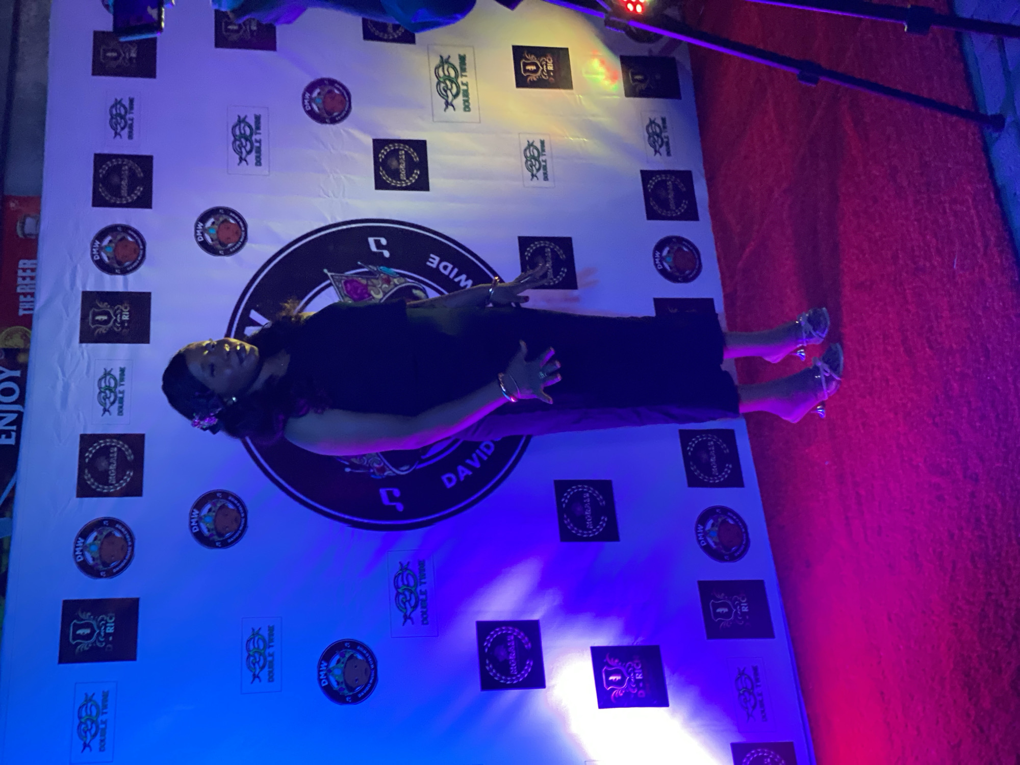 Here is a description of the image:
A woman poses for a photo in front of a backdrop decorated with logos and text, including "DOUBLE TWIN," "D-RICH," "MORALES," and "DMW WORLDWIDE." She is wearing a dark dress and heels. The lighting appears to be a mix of blue, red, and yellow. The location is in Iranla, Nigeria.