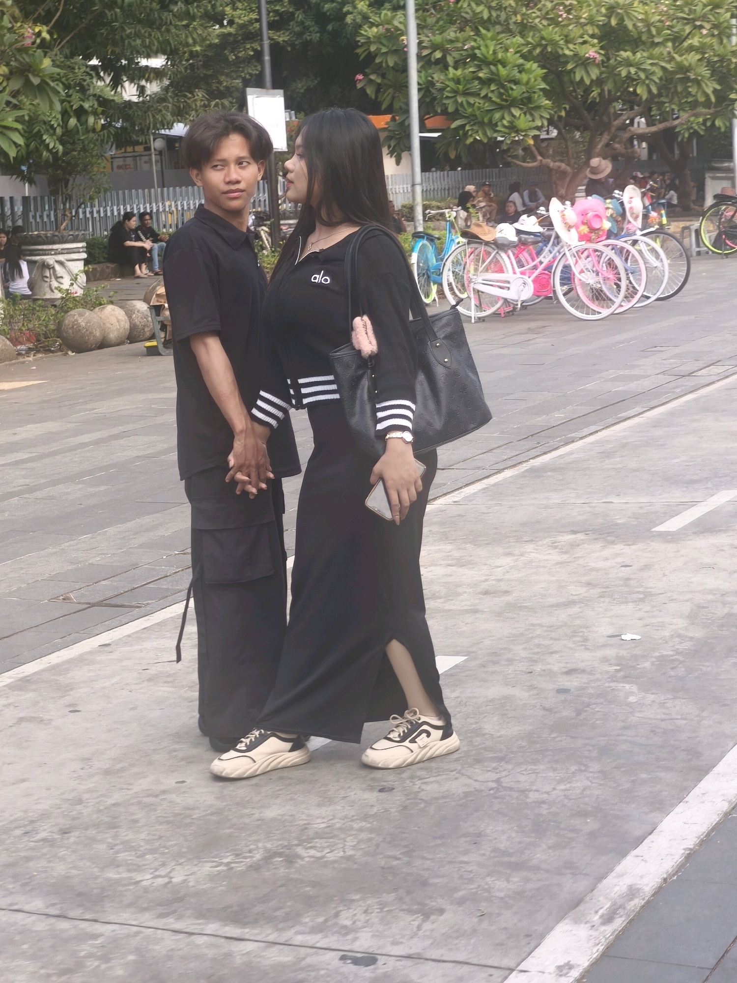 In West Jakarta, Indonesia, a young man and woman stand on a paved pathway, holding hands. The man, wearing a black polo shirt and black cargo pants, looks to his right, away from the camera. The woman, in a black long-sleeved shirt and a long black skirt, looks towards the man, her right hand holding a cell phone. A black tote bag hangs from her left shoulder. Behind them, a row of decorative bicycles, some painted pink and white with floral decorations, are parked. Trees and other people are visible in the background, suggesting an outdoor public space.