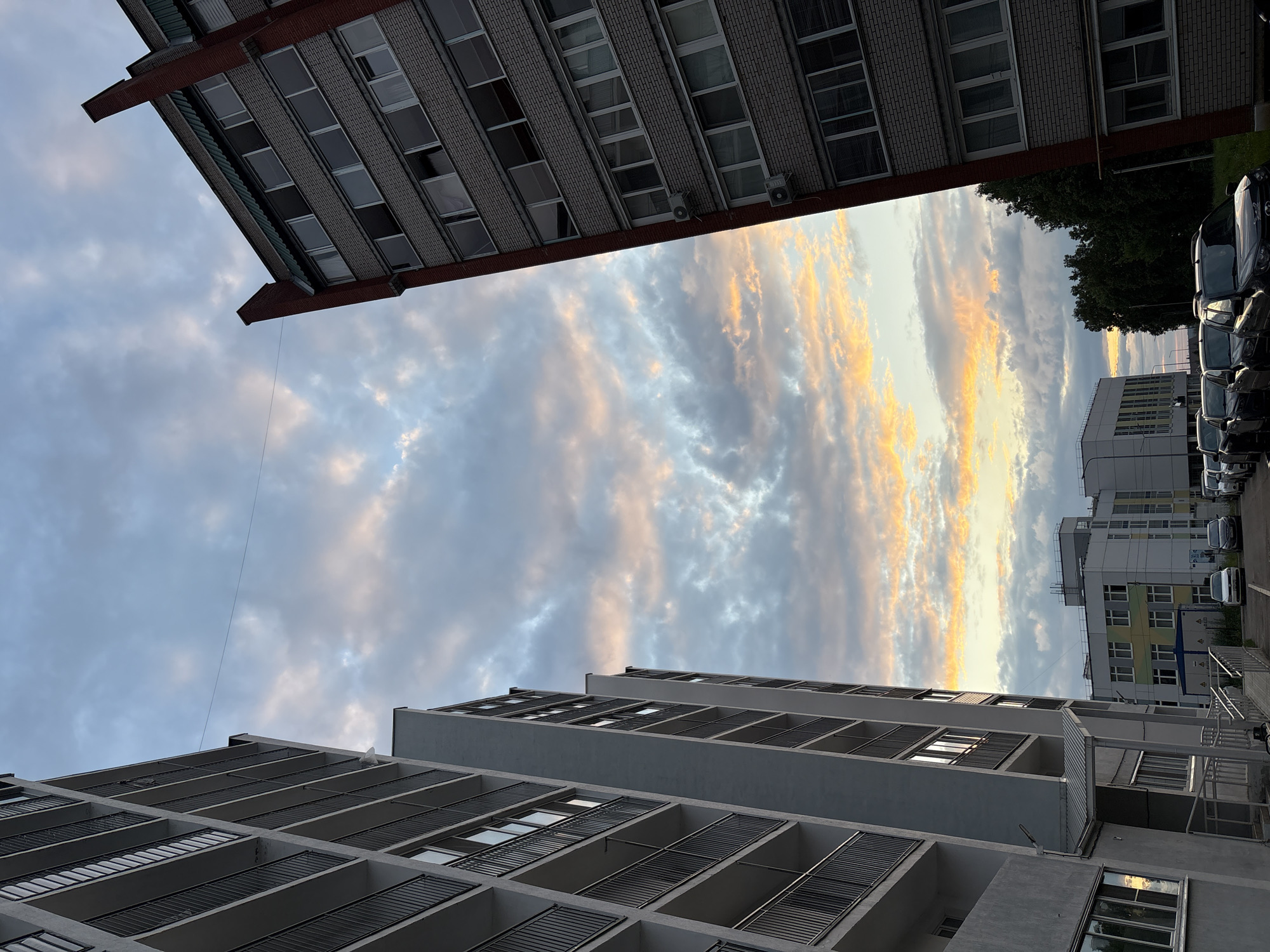 The image depicts a city scene in Chaikovsky, Russia, likely during sunset or sunrise, given the warm hues in the sky. Two tall apartment buildings frame the view, one on the left with a white facade and balconies with black railings, and another on the right with a brick exterior and red accents. The sky is filled with dramatic clouds, tinged with orange and yellow, contrasting with the cooler blues and grays. Below the buildings, a street is visible with several cars parked, suggesting a residential area. In the background, more buildings and trees are present. The perspective is from ground level looking upwards, emphasizing the height of the buildings and the expansive sky.