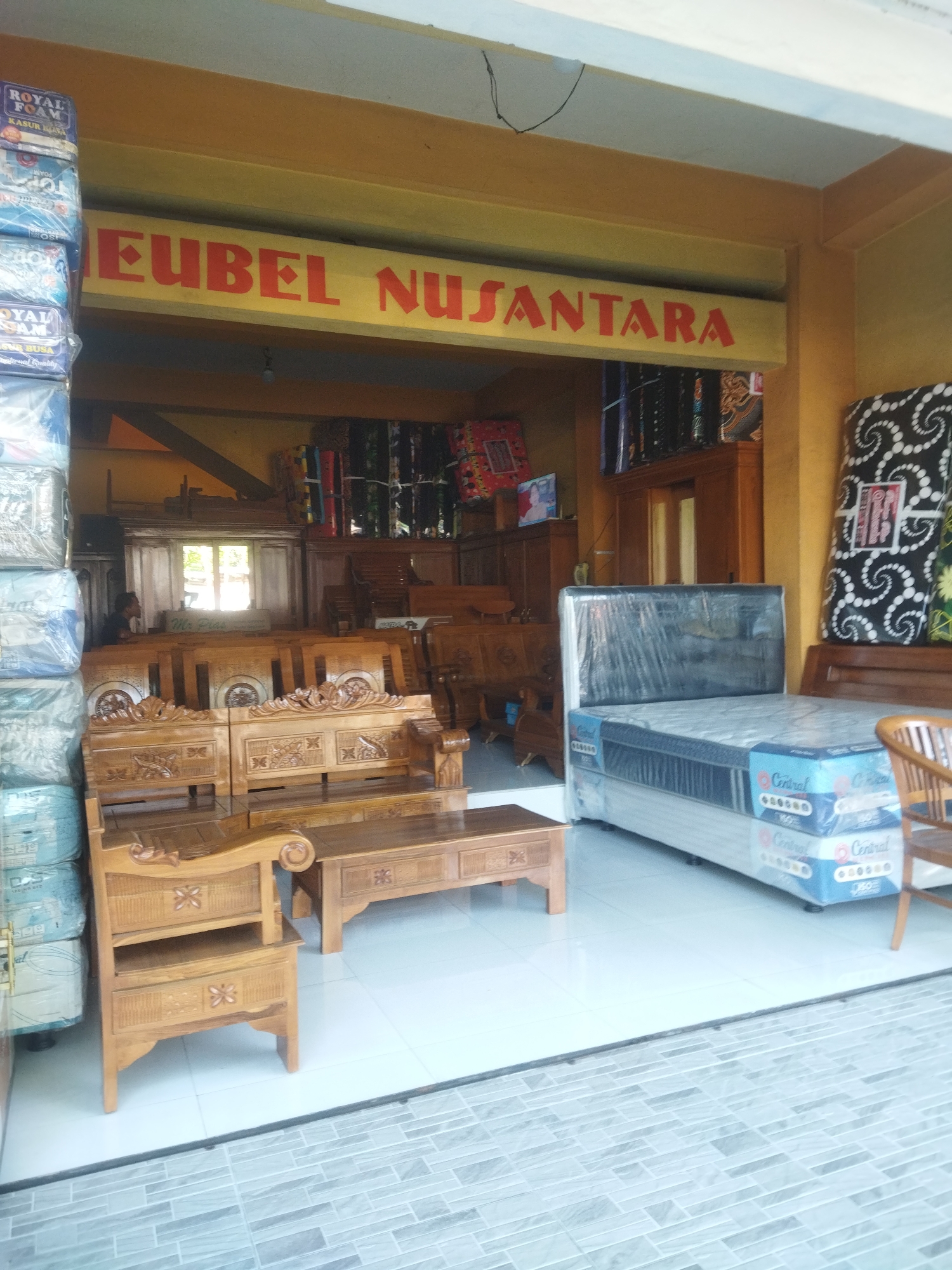 This is a medium shot of the exterior of a furniture store in Sukoharjo, Indonesia, with the store name "EUBEL NUSANTARA" prominently displayed above the entrance in red lettering on a yellow background. The store appears to be open, with a selection of furniture visible both inside and just outside the entrance.
In the foreground, there is a large wooden sofa with intricate carvings. To the right of the sofa, a bed with a mattress is displayed. The mattress is covered in plastic wrap and has "Central" branding visible. Further to the right, a wooden chair can be seen.
Inside the store, stacks of mattresses wrapped in plastic are visible on the left side, with "ROYAL FOAM" branding on some of them. Behind the sofa, several wooden cabinets and a large mirror are arranged. In the background, more furniture and stacked mattresses are visible. A person is partially visible inside the store, sitting and looking towards the interior.
The store has an open-air facade, with a tiled floor inside and a grey tiled area outside. The walls are painted yellow and white, and the ceiling is also yellow. The overall impression is of a retail environment selling mattresses and wooden furniture.