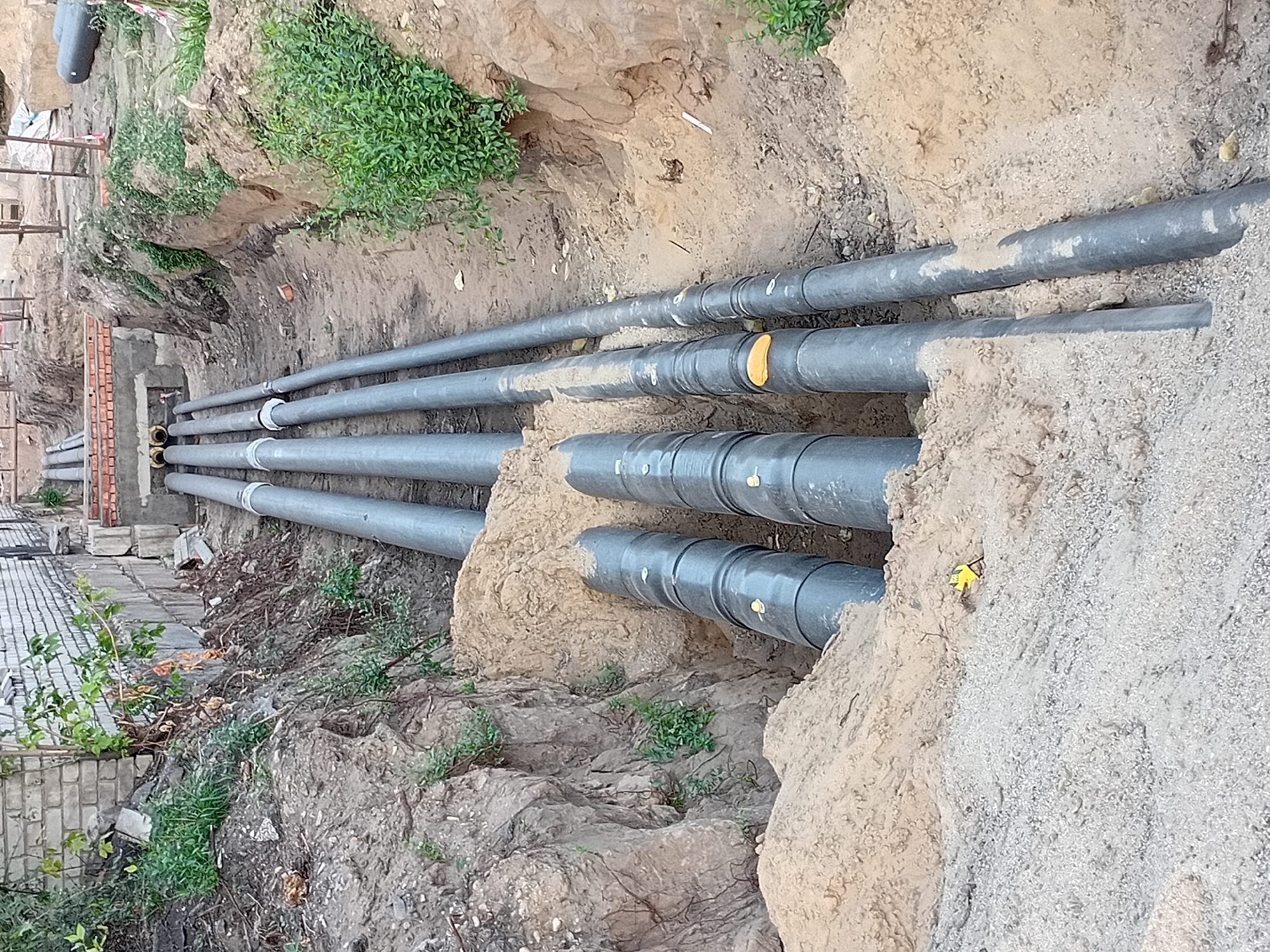 The image depicts a construction site in Semey, Kazakhstan, where a trench has been dug to lay a series of large, gray pipes. The trench walls are made of exposed soil and rock. Several pipes, appearing to be made of plastic or concrete, are visible, some partially buried in sand. One section of the trench shows a concrete structure with multiple openings where pipes enter. The surrounding environment is characterized by dirt, sand, and sparse vegetation. There are no people visible in the image. The scene suggests ongoing infrastructure work, possibly for water or sewage systems.