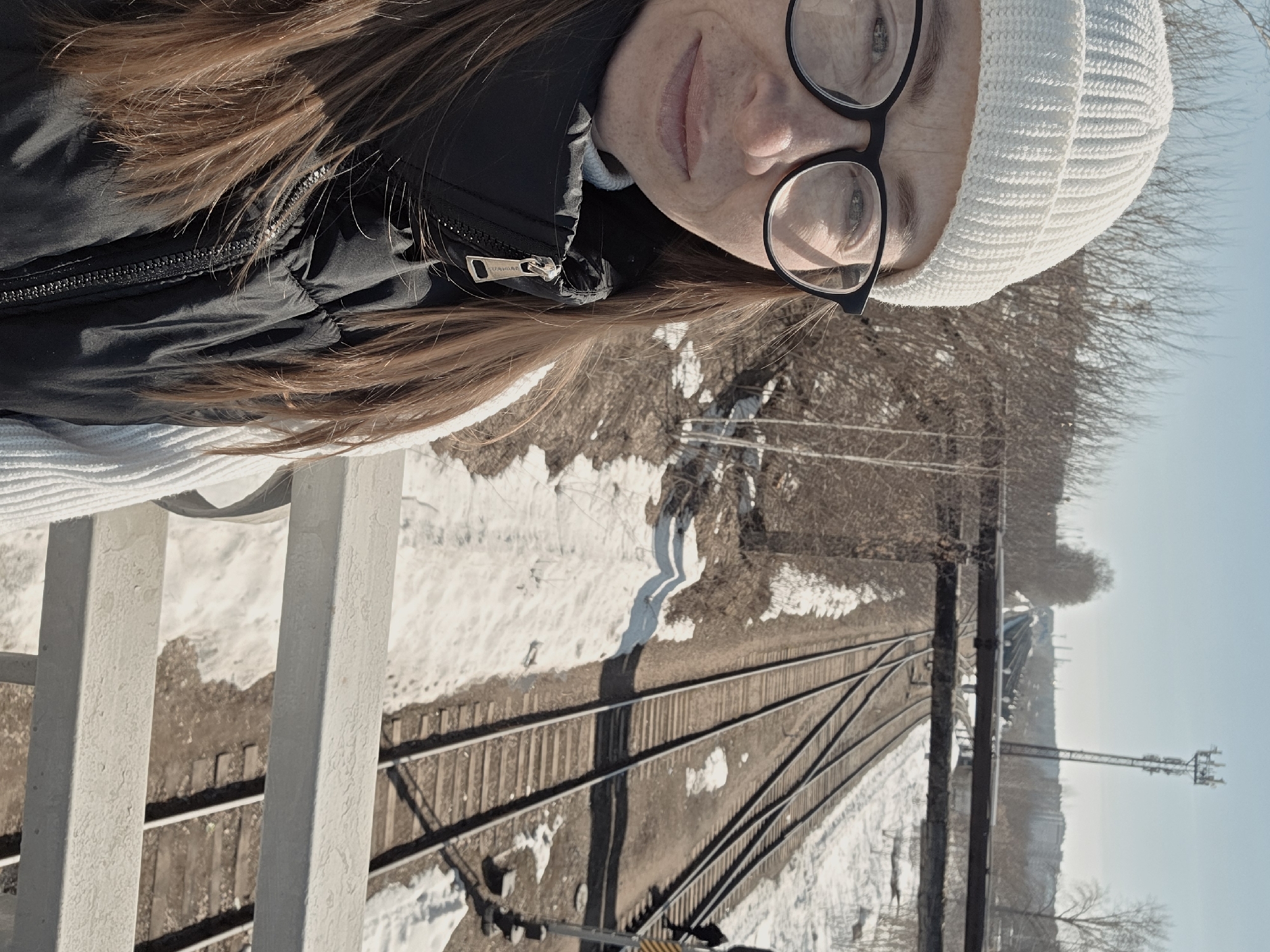 A photograph taken in Chaikovsky, Russia, shows a woman standing on a bridge overlooking railroad tracks. The woman has light brown hair, wears glasses, and is wearing a white beanie and a black jacket. The bridge is constructed of white painted metal. Below the bridge are railroad tracks. There is snow on the ground alongside the tracks. Bare trees and utility poles are visible in the background. The sky is light blue.