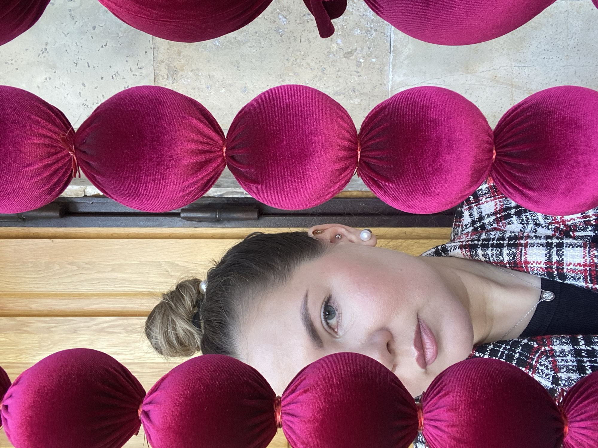 A woman is lying down with her face visible between rows of crimson velvet spheres that are tightly connected. The woman has fair skin, light-colored eyes, and her hair is pulled back into a bun. She is wearing a black and white patterned jacket with a dark top underneath, and a silver necklace is visible around her neck. Her ear is visible, adorned with multiple piercings and a pearl earring. The velvet spheres are connected by thin threads and appear to be part of a decorative element. The background consists of wooden panels and a stone surface. The photo was taken in Izhevsk, Russia.