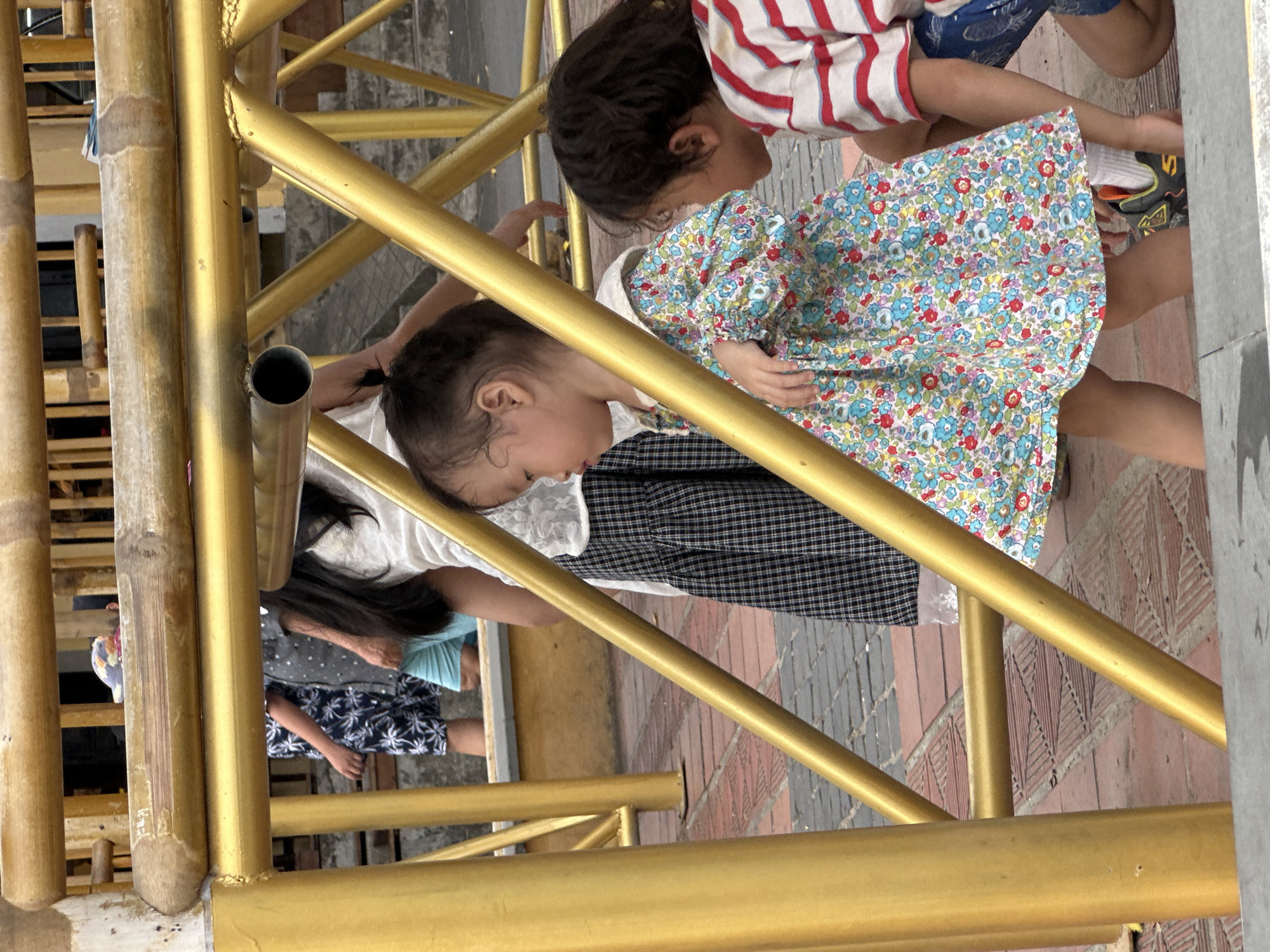 The image shows a group of children in an outdoor setting, likely a walkway or recreational area, in Hội An Ward, Vietnam. The children are interacting around a golden metal structure, possibly part of a playground or a decorative fence.
The foreground is dominated by the golden metal bars, some of which are cylindrical, and a bamboo pole. In the middle ground, a young child wearing a white top and a colorful floral dress is leaning forward, looking down. Next to this child is another child wearing a red and white striped shirt and blue shorts, also looking downwards. There is a third child partially visible in the background, wearing a blue and white patterned top and shorts.
The ground beneath them appears to be paved with bricks in a decorative pattern. The overall lighting suggests it is daytime, with no indication of specific weather conditions. The children's posture and focus suggest they are engaged in an activity together, perhaps playing a game or observing something on the ground. There is no visible text in the image. The scene conveys a sense of youthful energy and interaction.