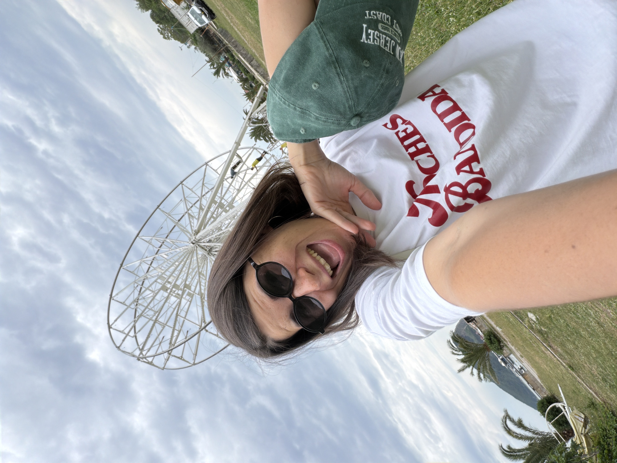 A medium shot shows a woman lying down and taking a selfie while holding a green New Jersey baseball cap above her head. The woman has shoulder-length graying brown hair and is wearing sunglasses and a white t-shirt with red cursive writing. She is sticking her tongue out and smiling. Behind the woman is a large Ferris wheel and a cloudy sky. In the distance, there is a body of water, a shoreline, green foliage, and buildings. The location is likely Budva, Montenegro, based on the shirt and Ferris wheel.