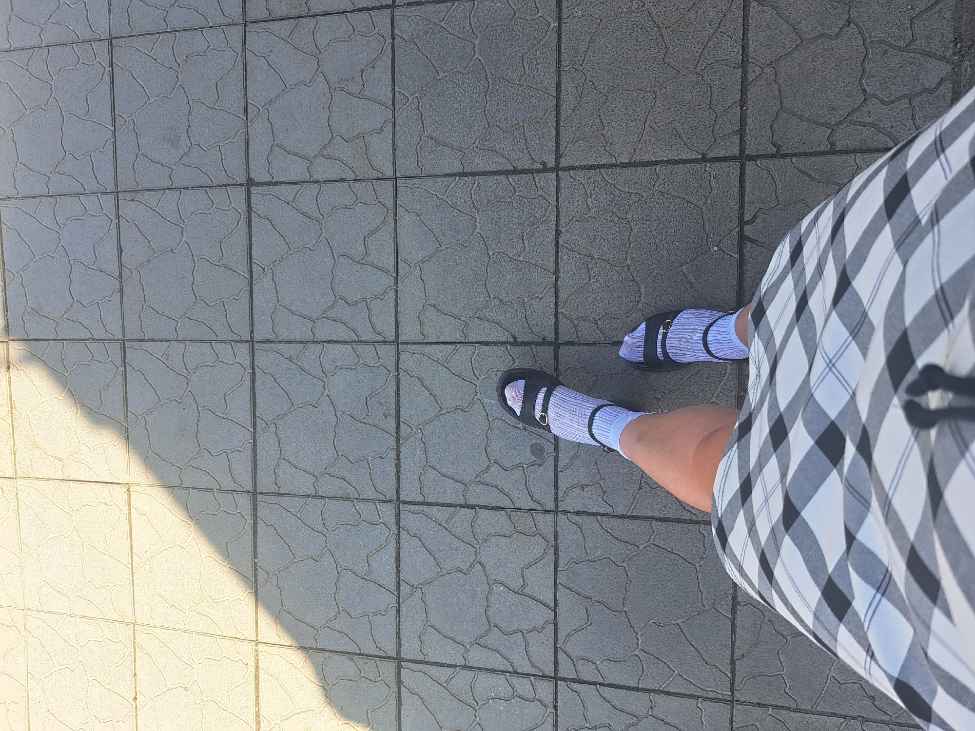 The image shows a first-person perspective of someone standing on a paved outdoor area. The ground is covered in square tiles with a textured, irregular pattern. A diagonal shadow casts across the tiles from the upper left to the lower right of the frame. The person is wearing a black and white checkered skirt, white socks with thin blue stripes, and black sandals. The scene appears to be a public walkway or sidewalk in Leningradskaya, Russia, likely on a sunny day given the distinct shadow.