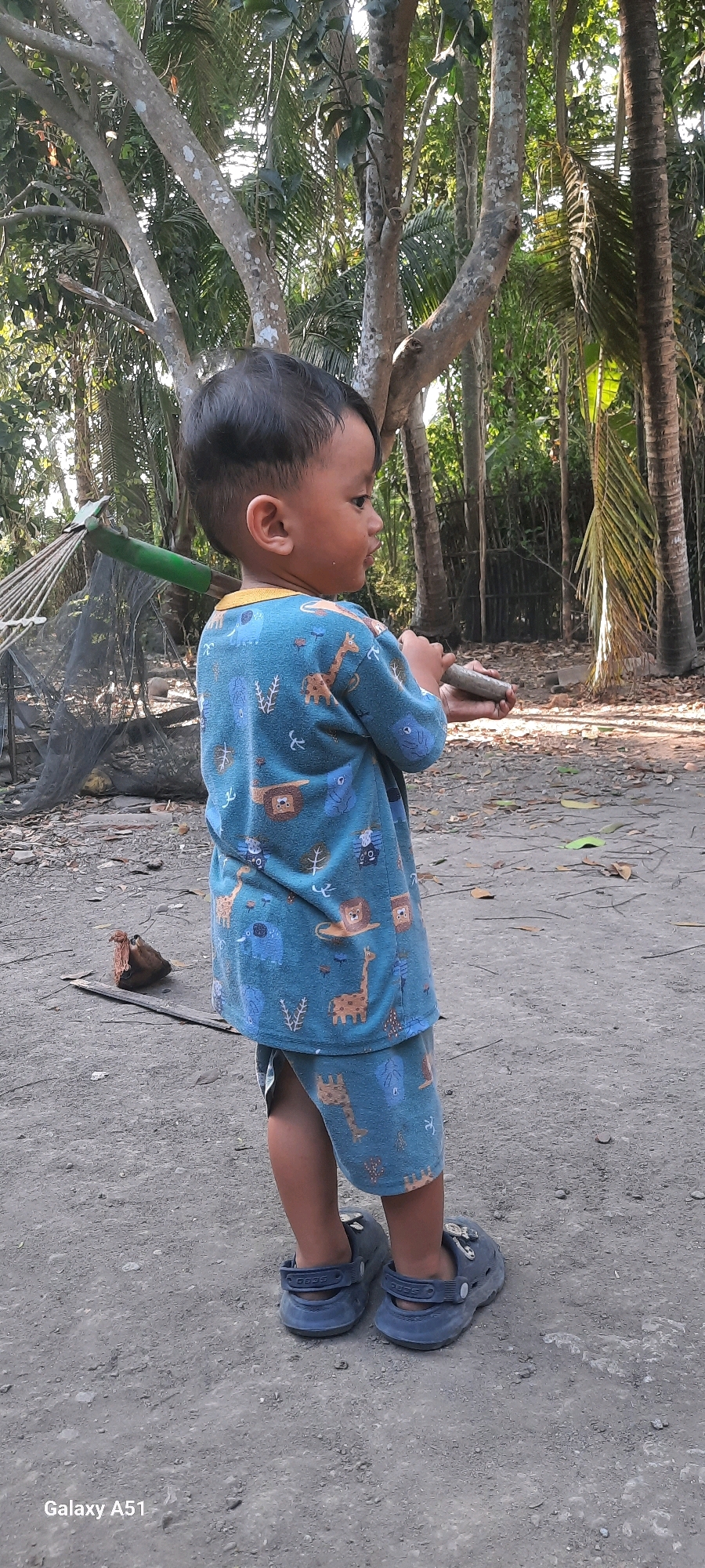 A young boy stands on a dirt path, looking to his right. He is wearing a blue pajama set with a safari animal print and blue Croc-style shoes. The boy is holding a small, cylindrical object in his hands. To his left, a rake with a green handle and a black net is leaning against a tree. The background consists of trees and foliage, suggesting an outdoor, possibly rural, setting. The image is geotagged to Jatitengah, Indonesia.