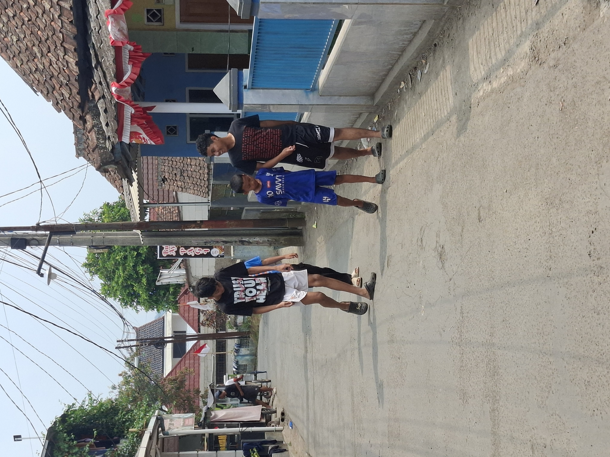 In the city of Karangtengah, Indonesia, on a bright day, three young males walk along a paved street in a residential area. The street is lined with simple houses, some with tiled roofs and one painted blue with red and white decorations. Electrical wires crisscross overhead. The first male, wearing a black t-shirt with white lettering and white shorts, walks with his head down. Behind him, a younger male in a blue t-shirt and blue shorts walks with his arm around the shoulder of the first. To the right of these two, a third, older male in a black t-shirt and black shorts walks slightly ahead, looking to his right and smiling. The ground is paved with asphalt and shows signs of wear and tear. The overall impression is of a casual stroll through a typical Indonesian neighborhood.