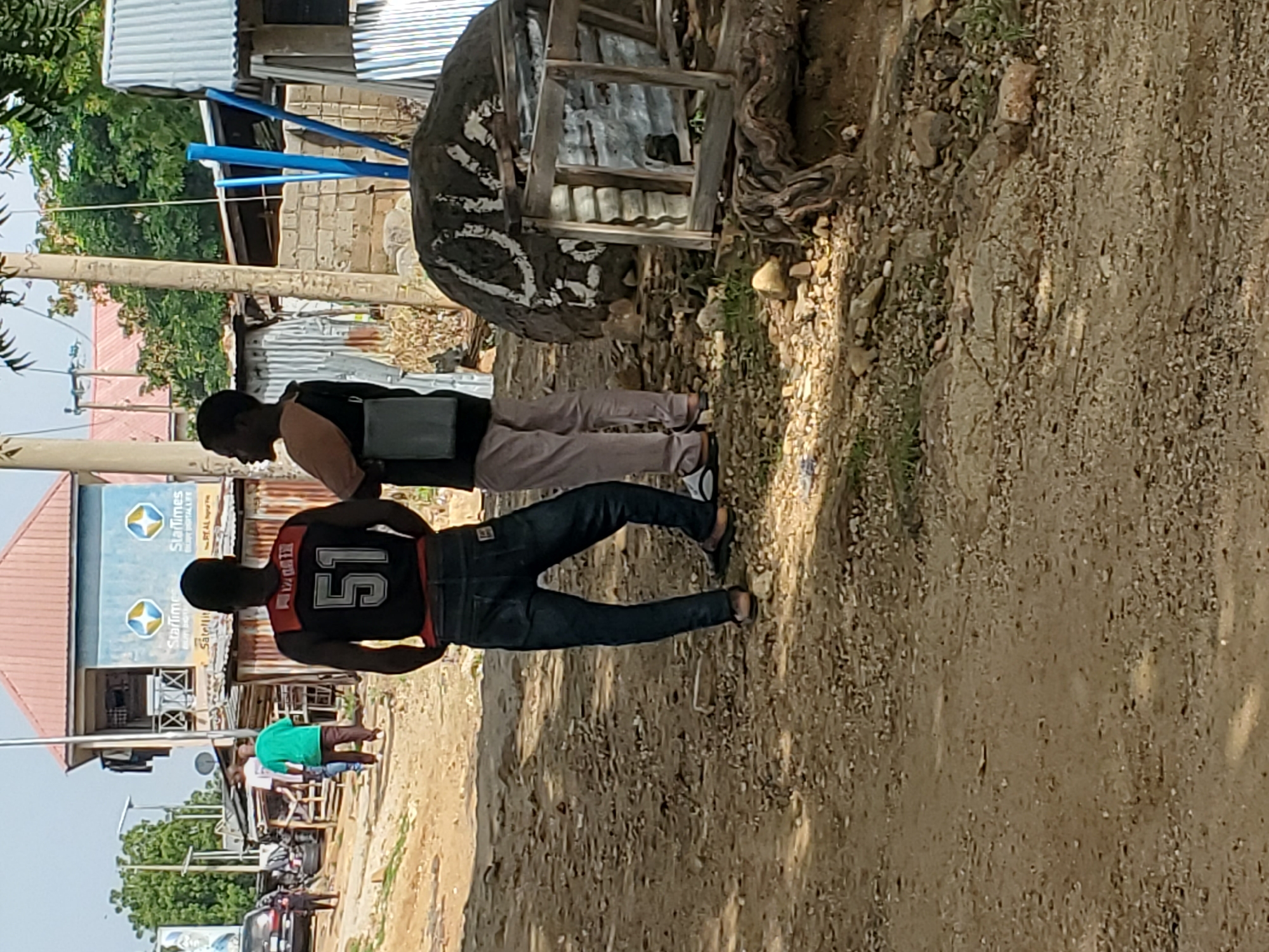 The image shows an outdoor scene with several people and objects. In the foreground, two men are walking on a dirt path. The man on the left is wearing a black shirt and light brown pants, and he has a black bag slung over his shoulder. The man on the right is wearing a black jersey with the number "51" on the back in white and red trim, and dark blue jeans. Behind them, other people are visible, including a person in a bright green shirt.
The background includes buildings with corrugated metal roofs, one of which has a blue sign with "StarTimes" logo. There is also a large, rough-textured object with some white markings, resembling a large rock or sculpture, placed near a wooden structure. The ground is covered with dirt, gravel, and some sparse vegetation. The overall impression is that of a somewhat dusty, rural or semi-urban environment.
The lighting suggests it is daytime, likely sunny, given the shadows. No specific weather conditions are evident beyond the dusty ground. The activity appears to be casual movement or walking along the path. The expressions of the people are not clearly discernible. Some text is visible on the "StarTimes" sign.