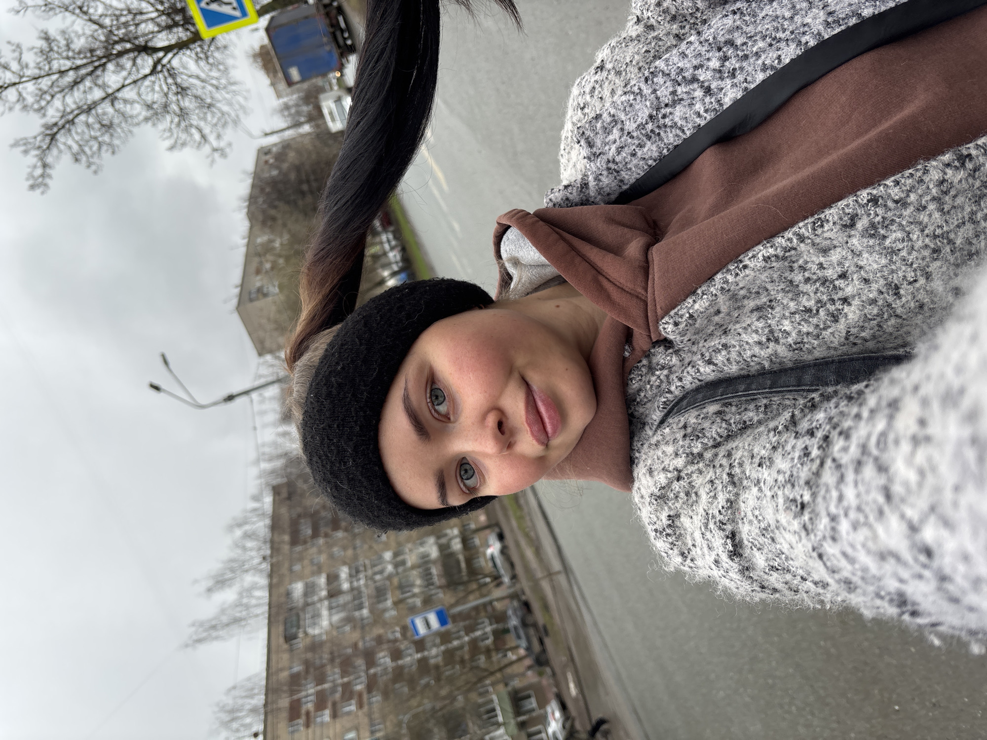 A selfie captures a young woman outdoors on an overcast day in Izhevsk, Russia. She is positioned in the foreground, looking directly at the camera with a calm expression and a slight smile. Her fair skin, light blue eyes, and eyebrows are prominent, framed by a black knitted headband. Her dark hair, with lighter tips, is pulled back into a high ponytail, part of which is visible extending out of the frame. She wears a brown hooded sweatshirt layered under a textured grey and white speckled coat or cardigan, with a black strap visible across her shoulder.
The background, tilted due to the selfie's angle, depicts an urban street scene. Dominating the upper left of the background is a bare-branched tree, indicating a colder season. Further back, multi-story residential buildings with multiple windows line the street. Several vehicles are parked along the road, which appears to be asphalt.
Visible street signs include a yellow square sign with a blue pedestrian crossing symbol and a blue rectangular sign featuring a white arrow pointing right. The sky is uniformly grey and overcast, suggesting daytime with no direct sunlight. The overall setting is consistent with a typical city street in a European or Russian urban area. No specific readable text is clearly visible on any of the buildings.