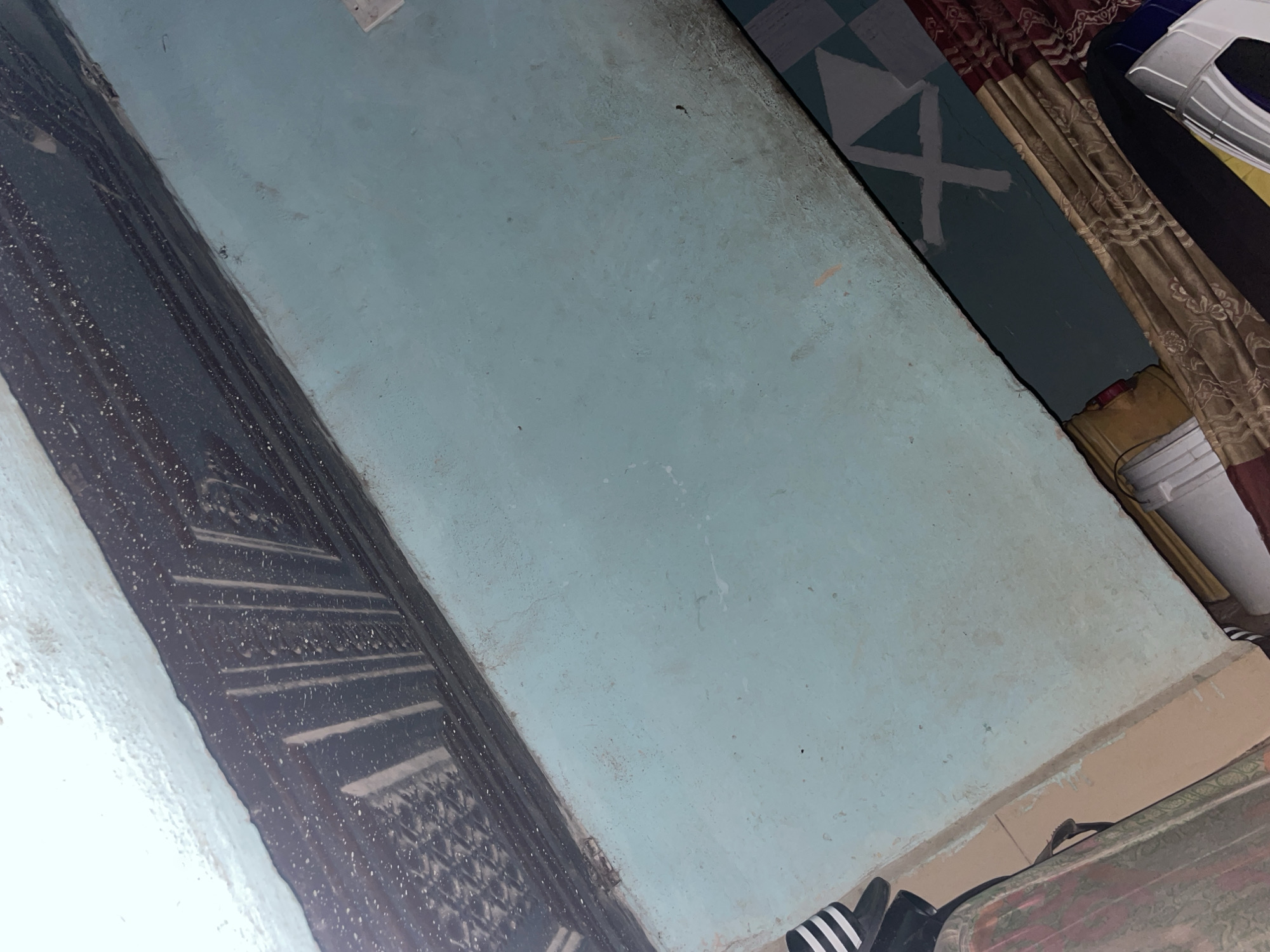 The image shows an interior scene, likely a room, with a focus on a surface that appears to be a table or shelf. The surface is painted a pale blue color and shows signs of wear, with some dirt and scuff marks visible. Along the top edge of this surface, there is a darker colored area with a textured pattern that looks like a decorative trim or perhaps a built-in element. White specks are scattered across this darker area, possibly dust or paint splatter.
Below the pale blue surface, there is a strip of material with a patterned design. Part of this pattern consists of dark blue and light gray squares, and on one of these squares, a white shape resembling a cross or an 'X' has been painted. Further down, there are draped fabrics in various colors, including red with a floral pattern and a golden brown material with an intricate design.
On the left side of the image, the edge of a black and white striped object is visible, appearing to be footwear, possibly a sandal or flip-flop. A portion of a patterned bag or luggage with floral motifs is also present.
The overall impression is of a somewhat cluttered and lived-in space. The image appears to have been taken from a high angle, looking down at the surface. There are no people visible in the image.
Given the context of Jalingo, Nigeria, this scene could represent a domestic interior, such as a bedroom or living area, or possibly a workspace. The type of materials and finishes suggest a typical residential setting within the city.