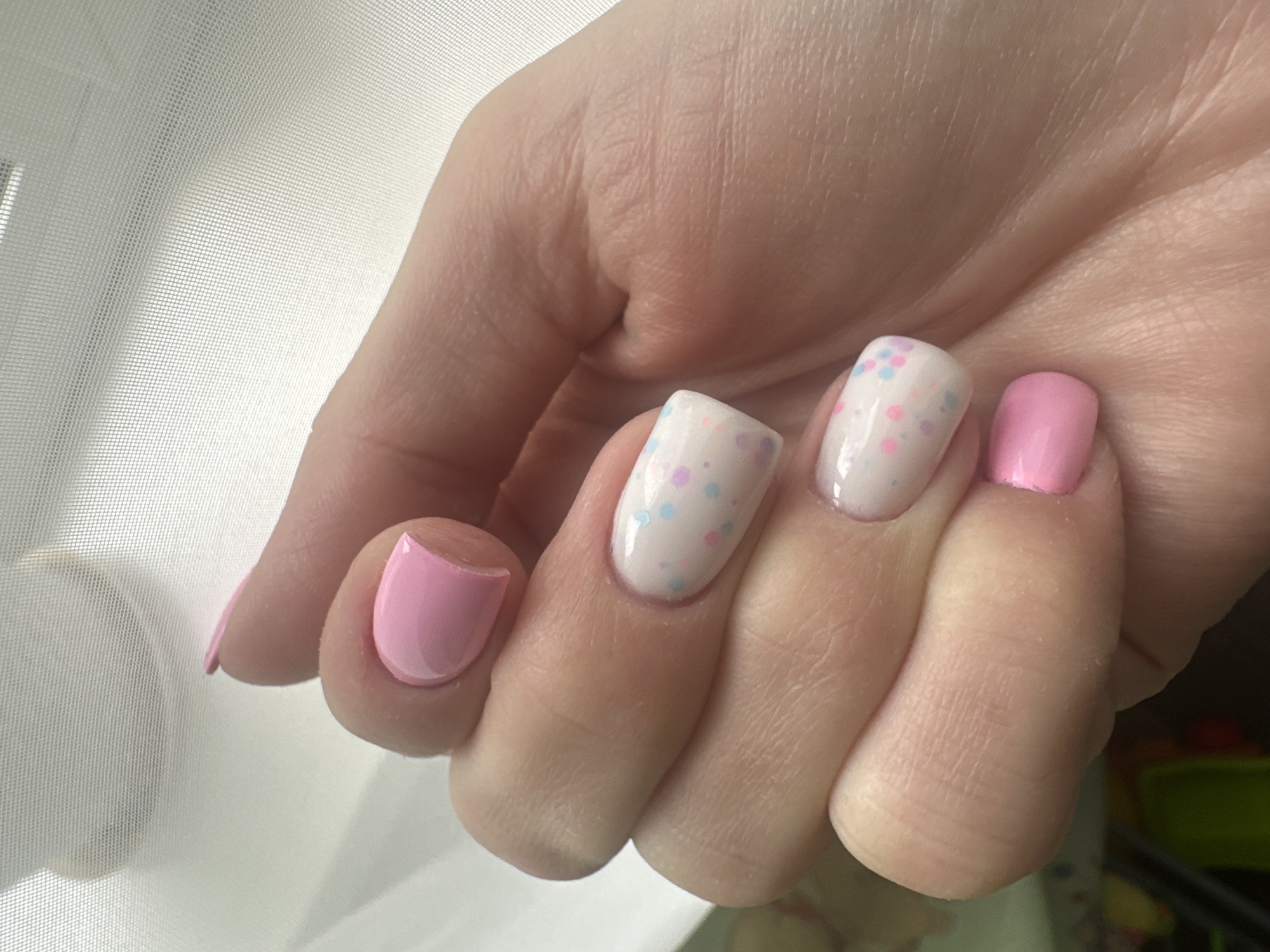 The image displays a close-up of a person's left hand, showcasing manicured nails. The nails on the thumb, index finger, and pinky finger are painted a solid pastel pink. The middle and ring fingers have a white base coat adorned with small, scattered pastel dots in pink, blue, and yellow. The hand is positioned against a translucent white or light-colored fabric, possibly a sheer curtain, with visible grid patterns. Natural light appears to be coming from the right, casting a soft illumination on the hand and creating subtle shadows. The background is out of focus and does not provide any discernible details about the location. There are no indications of the city of Sayanogorsk, Russia, or any other specific geographical information within the image.