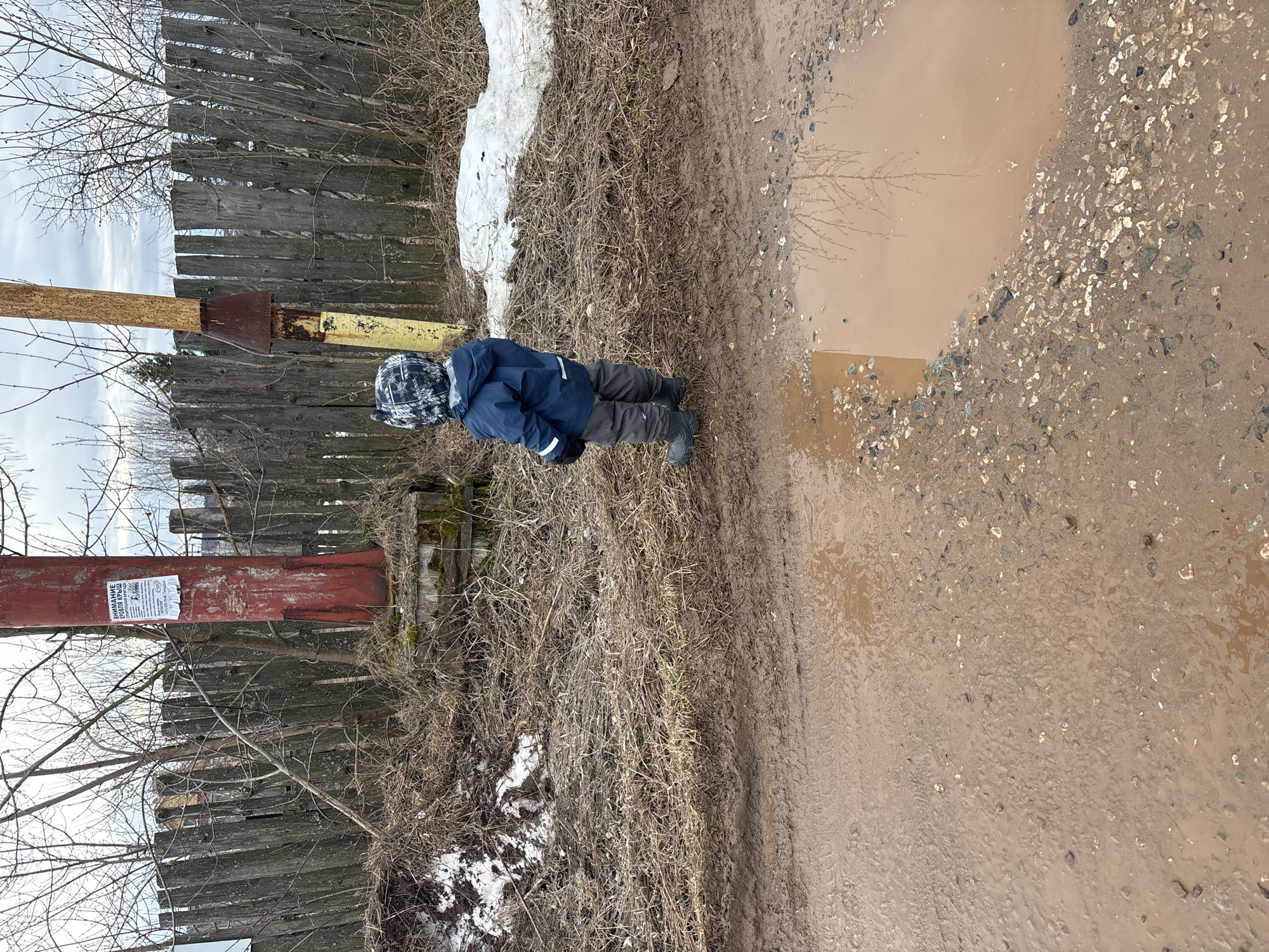 A small child wearing a blue jacket and patterned hat stands near a large muddy puddle in a rural area. The ground is covered in mud, dry grass, and some patches of snow. A dilapidated wooden fence runs along the left side of the scene, with a rusted metal post attached to it. A white sign is affixed to the post. The puddle reflects bare branches of a tree. The location appears to be a roadside in a neglected area. The city is Ferma, Russia.