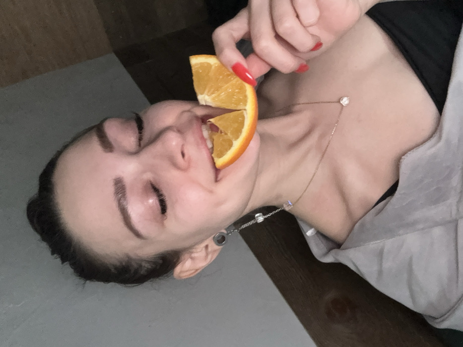 A female individual is depicted in an indoor setting within Reutov, Russia. Her face is in the upper half of the frame, with her eyes closed or partially closed and her mouth open, biting into a segmented orange. Her right hand holds the orange. She has dark hair pulled back, visible eyelashes, and shaped eyebrows. Her left earlobe features a plug or tunnel, from which a long, dangling earring with multiple clear, cube-shaped beads extends. A thin necklace with a small, clear pendant rests on her collarbone. Her fingernails are painted red. She wears a black garment underneath a light gray or muted-colored outer garment draped over her shoulders. The background is dim and indistinct, with a vertical wooden element visible to the right and a solid-colored wall to the left. The lighting is low, illuminating the subject primarily from the front.