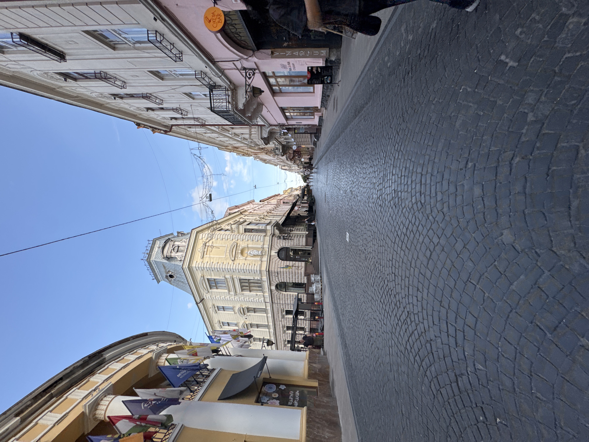 This image depicts a street scene in Chernivtsi, Ukraine, likely a central European city with historic architecture. The foreground is dominated by a cobblestone street, curving gently into the distance. On the left side of the street, a curved yellow building with white columns and decorative flags is visible. Adjacent to it, a darker awning covers an entrance with a chalkboard sign displaying text in Ukrainian.
Further down the street, on the left, a prominent cream-colored building with a distinctive mansard roof and ornate architectural details stands at a corner. This building appears to be a landmark.
The right side of the street features a row of multi-story buildings with a light pink facade in the foreground, housing shops or cafes. A circular orange sign with the text "CIKEPA BAR" is prominently displayed. Further down, buildings with lighter facades and balconies line the street. Several people are walking or standing along the sidewalks and in the street. Overhead, string lights are visible, suggesting a festive or commercial atmosphere. The sky is a clear blue with a few wispy clouds. The overall impression is of a vibrant, historic urban environment.