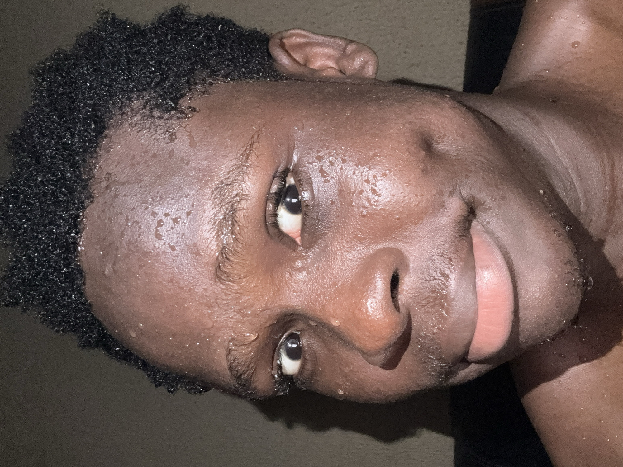 A close-up, tilted portrait of a person with dark skin and short, dark, textured hair. The person is sweating, with droplets visible on their face and upper body. Their eyes are open and looking slightly upward. The background is dark and indistinct. There is no contextual information to determine the location within Onitsha, Nigeria.