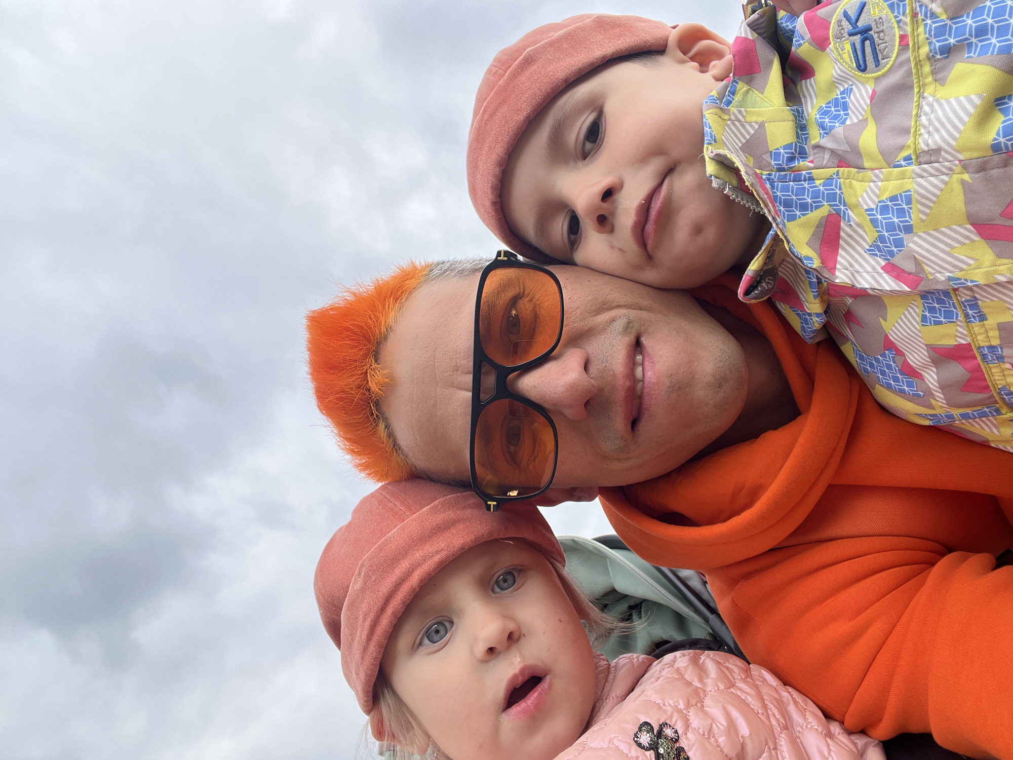 Here is a detailed description of the image:
The image is a low-angle selfie of a man with bright orange hair, wearing orange-tinted sunglasses and an orange hoodie. He is smiling, and his face is positioned in the center of the frame.
A young boy, wearing a salmon-colored beanie and a patterned jacket with the letters "SK for KIDS" on it, is resting his head on the man's head.
A toddler girl, wearing a salmon-colored beanie and a light pink quilted jacket, is positioned below the man. She is looking up at the camera with an open mouth.
The background is a cloudy sky.
Based on the presence of "SK for KIDS" branding, it is possible that the photo was taken in or near Chaikovsky, Russia, but this is a conjecture based on the branding, not definite proof of the location.