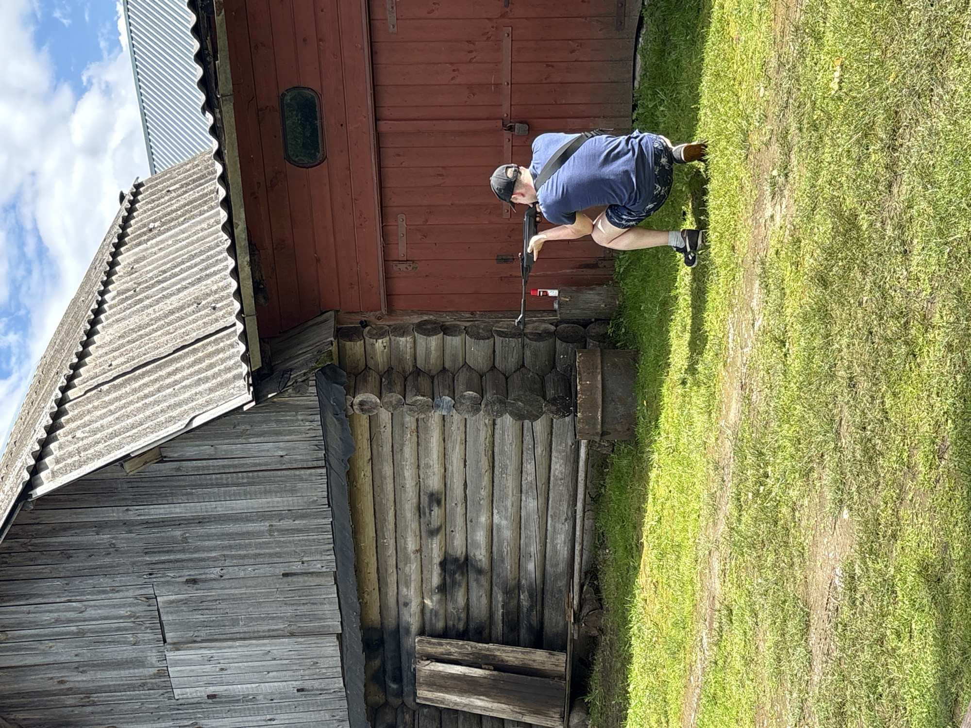 The image depicts a person crouching in front of a rustic wooden building, holding a rifle. The person is wearing a blue t-shirt, dark shorts with a pattern, a black baseball cap, and sneakers. They are aiming the rifle at something out of frame. The building appears to be made of weathered logs and has a reddish-brown wooden section with an oval window. Next to it is another section of the building with grey, corrugated roofing and wooden siding. The scene is set outdoors on a sunny day, with green grass covering the ground. The person's stance and the rifle suggest an activity related to shooting or target practice. The overall atmosphere is casual and outdoorsy, set in a rural location. The location context provided is Дресвяново, Russia, which aligns with the rustic architecture and natural setting. There is no visible text in the image.