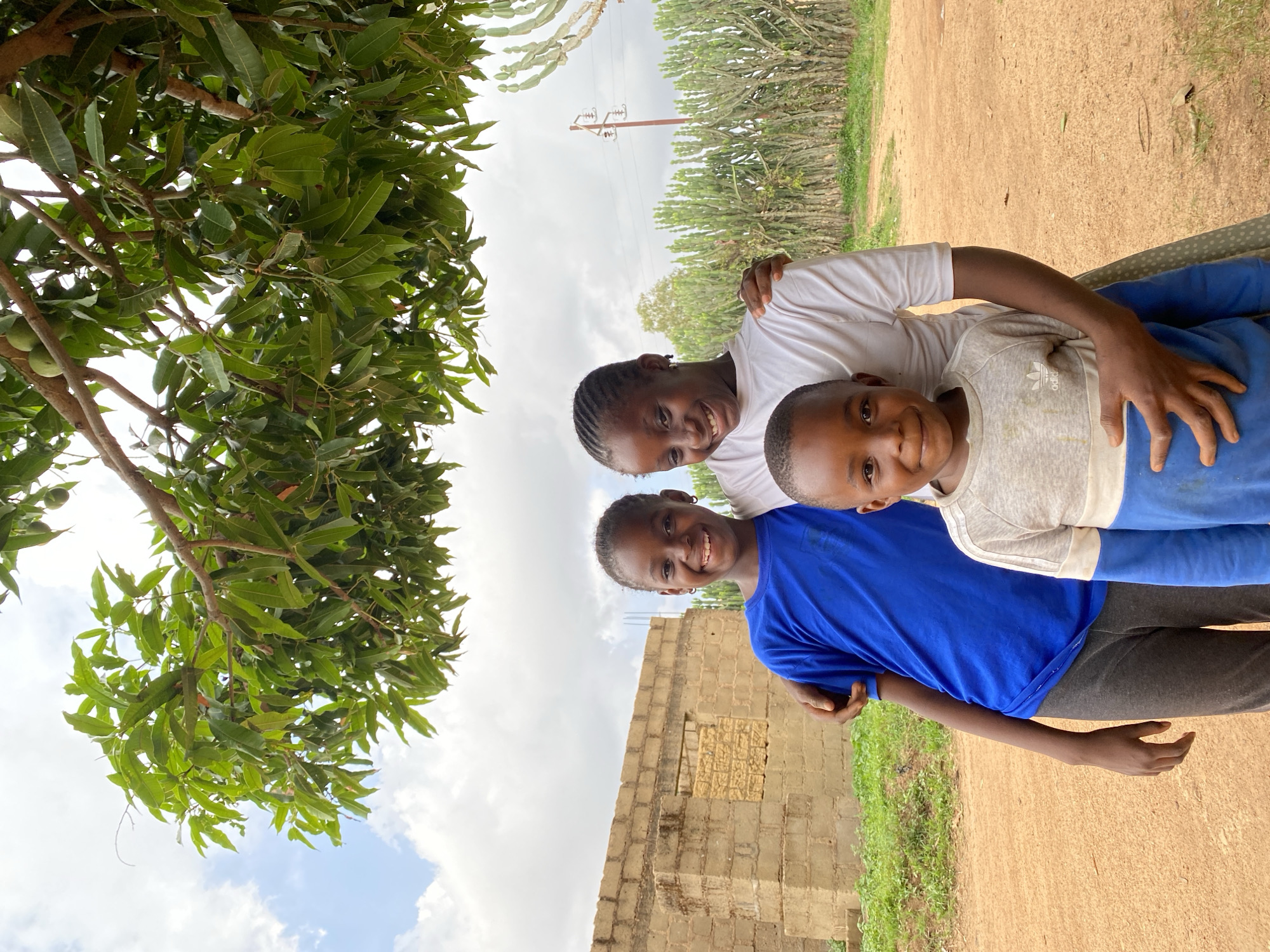 Three smiling African children pose closely together in an outdoor setting in Kuru Dungus, Nigeria. A girl with braided hair wears a white t-shirt and places her arm around the child in the center. The central child, a boy, wears a gray and white shirt. The child on the left wears a bright blue t-shirt. They appear to be standing next to a wall constructed of tan blocks. A large tree with green leaves partially obscures the left side of the frame. In the background, utility poles and tall, cactus-like plants are visible. The ground is a mix of dirt and grass. The sky is cloudy.
