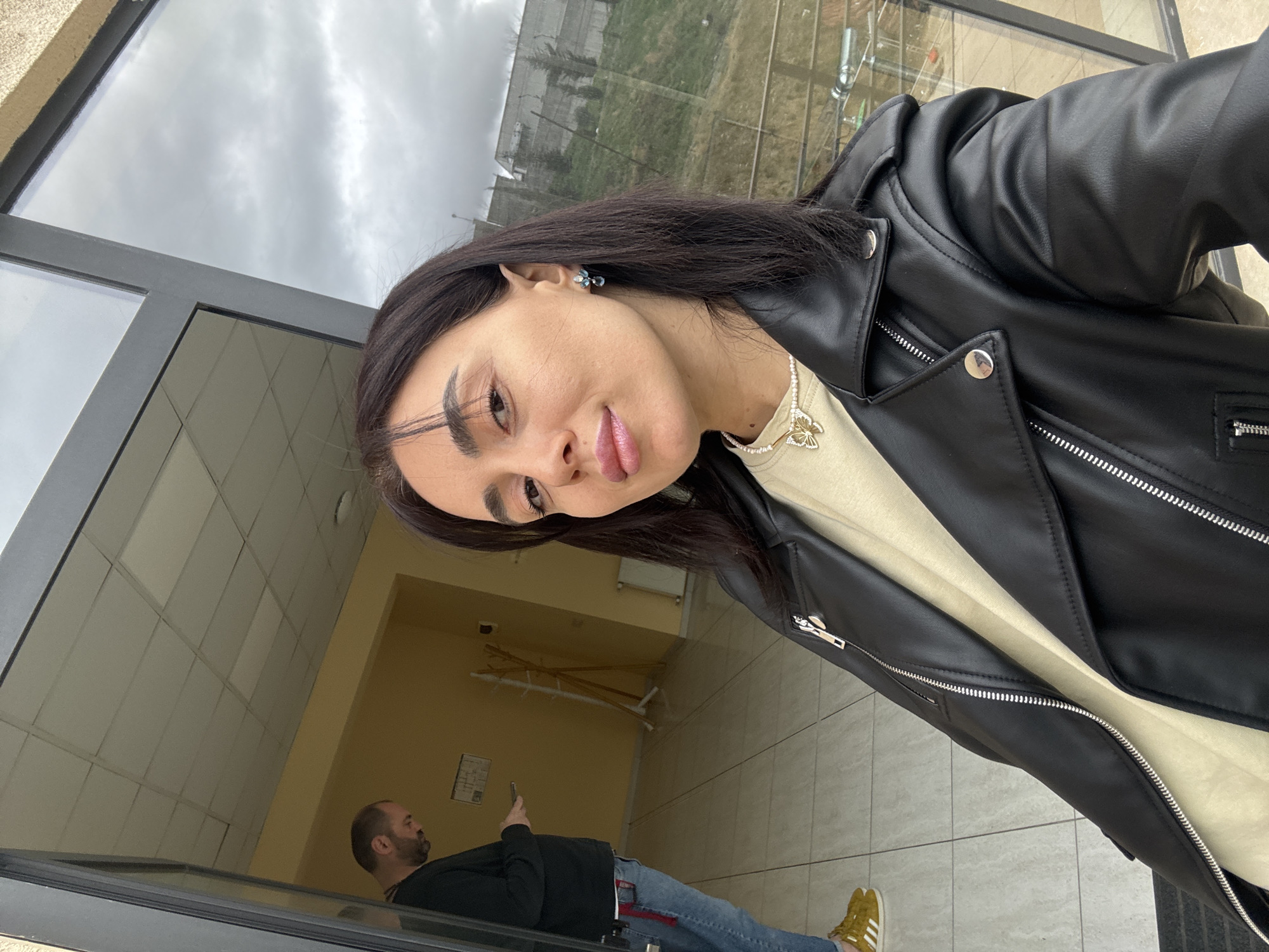The image displays a selfie-style shot of a female in the foreground, positioned in an indoor space with a large glass exterior. The female, in her late 20s or early 30s, has dark hair and wears a black faux leather jacket over a light-colored top. She is accessorized with a pearl necklace featuring a gold butterfly pendant and a blue and silver earring on her visible right ear. Her lips are a light pink color.
Behind her, to the left, a male is visible standing inside what appears to be a brightly lit room or hallway. He has a beard and is dressed in a black long-sleeved shirt, blue jeans with red fabric accents, and yellow Adidas sneakers. He is looking down at a smartphone. The indoor environment features light yellow walls, white tiled flooring, and a white suspended ceiling. A wooden coat rack stands against the far wall, and a white radiator is visible on the right wall. A document or sign is affixed to the left wall.
Through the large glass window or door to the right, an outdoor scene is reflected. The sky is overcast with gray clouds. A green, grassy hillside with scattered trees is visible, topped by distant buildings with reflective surfaces. This hilly urban landscape is consistent with areas within the city of Tbilisi, Georgia. The setting appears to be an entrance or waiting area of a building that offers a view of a neighboring hillside.