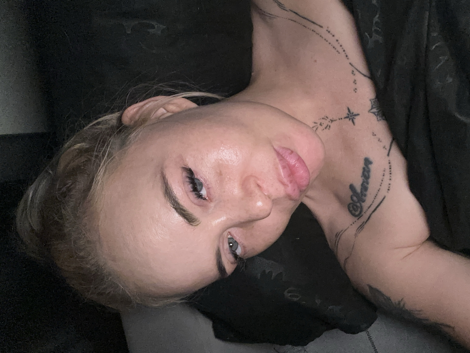 This image depicts an adult female, likely in her 20s-30s, captured in a close-up, dimly lit indoor setting in Unknown, Unknown. Her blondish-brown hair is gathered, with some wisps visible around her forehead. She possesses light blue eyes with prominent lash extensions and defined eyebrows. Her face is angled slightly towards the camera, and her lips are pursed in a pouting expression. The skin on the right side of her face (viewer's left) exhibits a shiny appearance. Multiple tattoos are visible on her upper torso: the word "Amor" in a script font is present on her right collarbone, surrounded by abstract lines and dots. Below this, on her neck/upper chest, is a linear tattoo composed of small decorative elements culminating in a star shape. A larger, dark, feathered or leaf-like tattoo is visible on her left upper arm/shoulder. She is partially covered by dark fabric, possibly bedding or a blanket. The background is predominantly dark and out of focus, with a dark pillow or cushion visible behind her head on the left side of the frame.