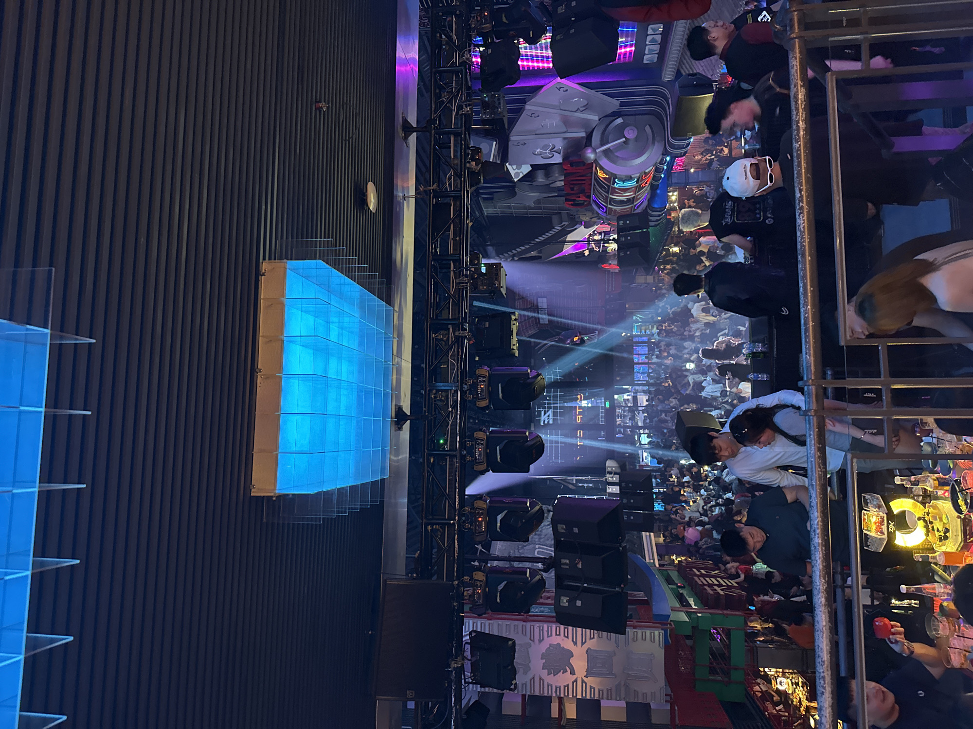 The image depicts a crowded indoor venue, likely a nightclub or entertainment establishment, located in the city of Thủ Đức, Vietnam. The atmosphere is illuminated by vibrant, colorful lights, including a large, rectangular blue lighting fixture suspended from the ceiling. The venue features a multi-level design with numerous patrons occupying various areas.
In the foreground, a metal railing separates the viewer from a group of individuals seated at tables laden with drinks and food. One person in the foreground is holding a red object. Further back, several people are standing and interacting. Notable figures include a man in a white shirt with his arm around a woman, and another man in a dark polo shirt.
The midground reveals a densely packed crowd of people standing and moving. Above them, stage lighting equipment, including spotlights and speakers, is mounted on trusses. Behind the crowd, a large, decorative structure resembling a casino slot machine with flashing neon lights is visible. This structure is adorned with playing card symbols, including spades and clubs.
The background shows more of the crowded venue, with additional lighting and what appears to be architectural elements. The overall impression is one of a lively and energetic nightlife scene.