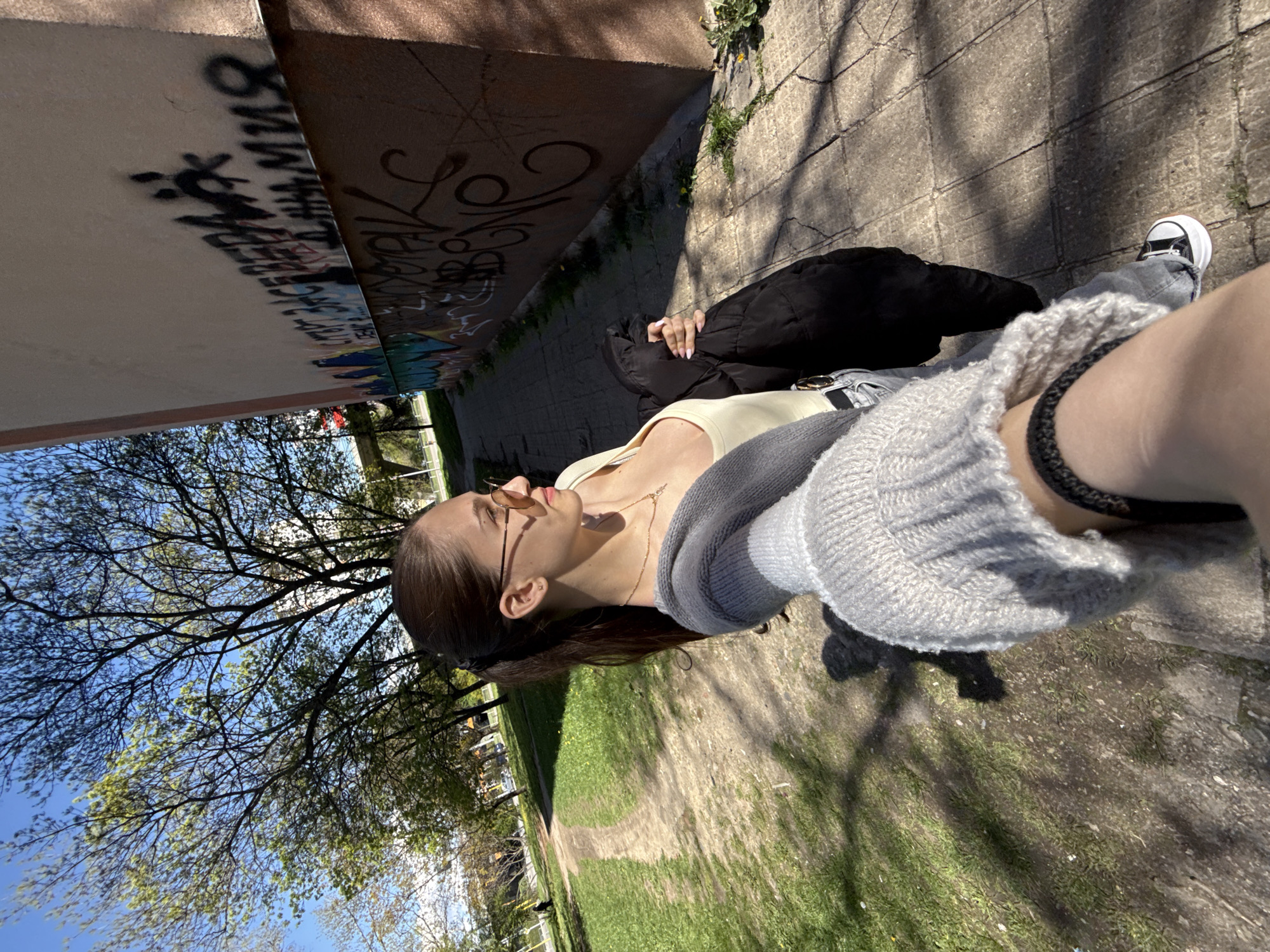 The image depicts a young woman in Sofia, Bulgaria, seated outdoors, with her legs and feet partially visible. She wears sunglasses, a light-colored top, a grey cardigan, and black accessories on her leg. Her clothing suggests a casual style.
The background features a mix of urban and natural elements. To the left is a building or structure covered in graffiti, indicating an urban setting. Beyond the building, a park or green space with trees and grass stretches out. The composition suggests the woman is sitting near the edge of an urban area transitioning into a park.
Given the presence of graffiti and the general urban-natural mix, the location is likely a park or green area in Sofia, Bulgaria, which is situated adjacent to buildings. The picture is taken during the daytime, with shadows and light visible across the scene.