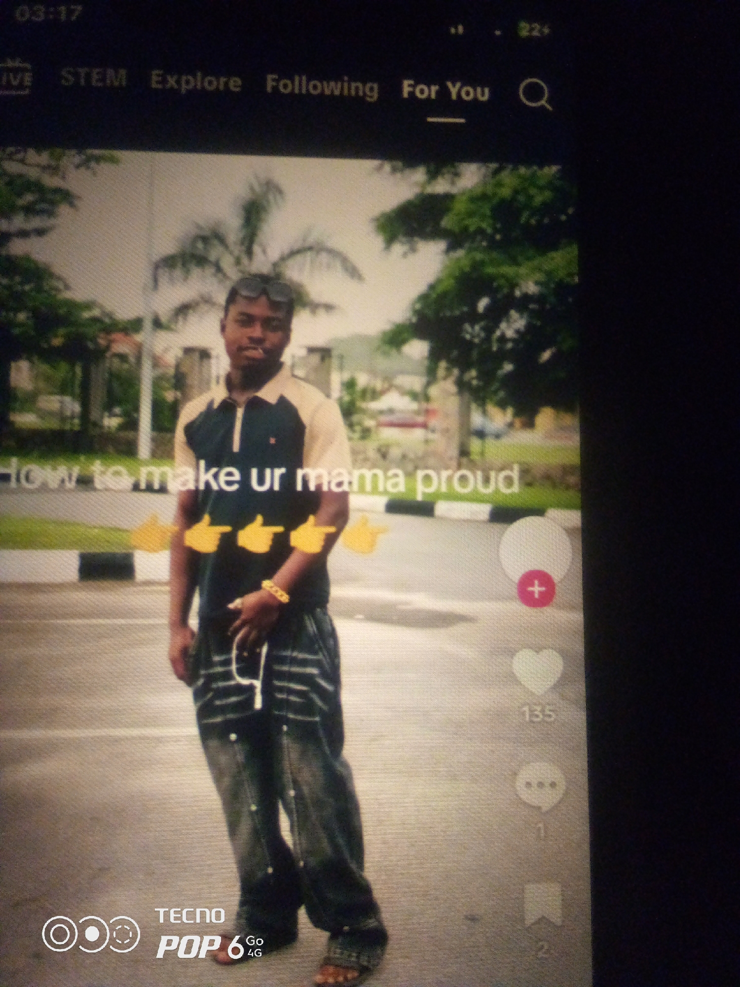 The image displays a TikTok-style video interface. The video features a young Black male standing outdoors, wearing a tan and navy polo shirt, baggy jeans, and a gold chain and bracelet. He has sunglasses perched on his head. The text overlay on the video reads "how to make ur mama proud". To the right of the video, social media interaction icons are visible, including a red plus sign, a white heart icon with the number "135" below it, a speech bubble icon with the number "1" below it, and a bookmark icon with the number "2" below it. The top of the screen shows the time "03:17" and navigation options like "LIVE", "STEM", "Explore", "Following", and "For You", along with a search icon. A "TECNO POP 6 Go 4G" logo is visible in the bottom left corner. The background appears to be an urban or suburban setting with trees and a road. While the image is presented as being in "Howayi, Nigeria", there is no visual information in the image itself to confirm this specific location.