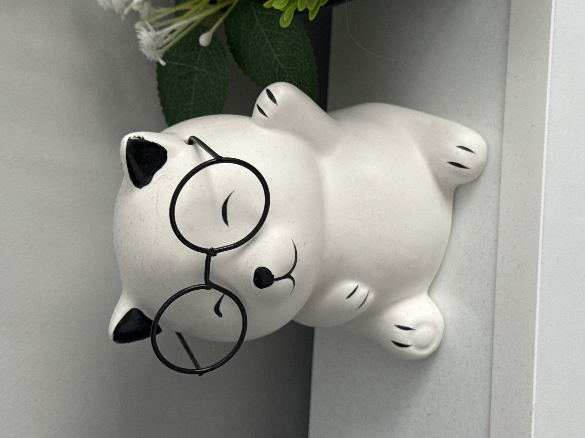 A white ceramic cat figurine wearing black wire-rimmed glasses lies on its back. The cat has black markings on its ears and paws, and a smiling mouth. A bouquet of white flowers with green leaves is partially visible above the cat's head. The figurine is placed on a white surface with a vertical white panel to the right, creating a corner. The location is Stara Zagora, Bulgaria.