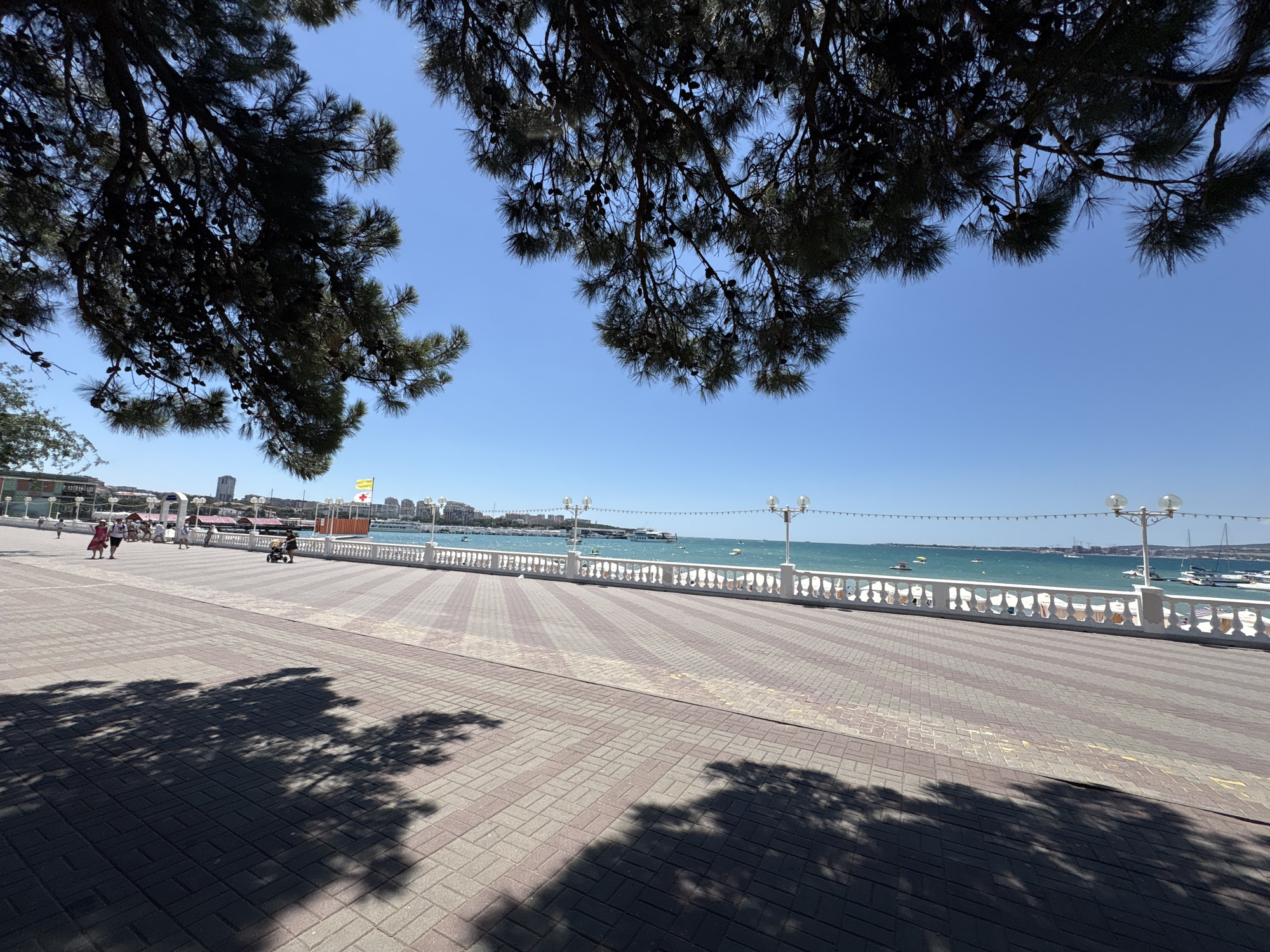 The image captures a sunlit promenade in Gelendzhik, Russia, likely during the daytime. The foreground is dominated by a patterned brick walkway, with dark tree shadows stretching across it. To the right, a white balustrade lines the edge of the promenade, separating it from the azure sea. Beyond the balustrade, several boats are visible on the water, and the coastline is dotted with buildings. People are present on the promenade, some walking, while others appear to be pushing a stroller or sitting. Overhead, the dark branches of pine trees, some with visible pinecones, frame the clear blue sky. The overall scene depicts a pleasant, recreational waterfront area.