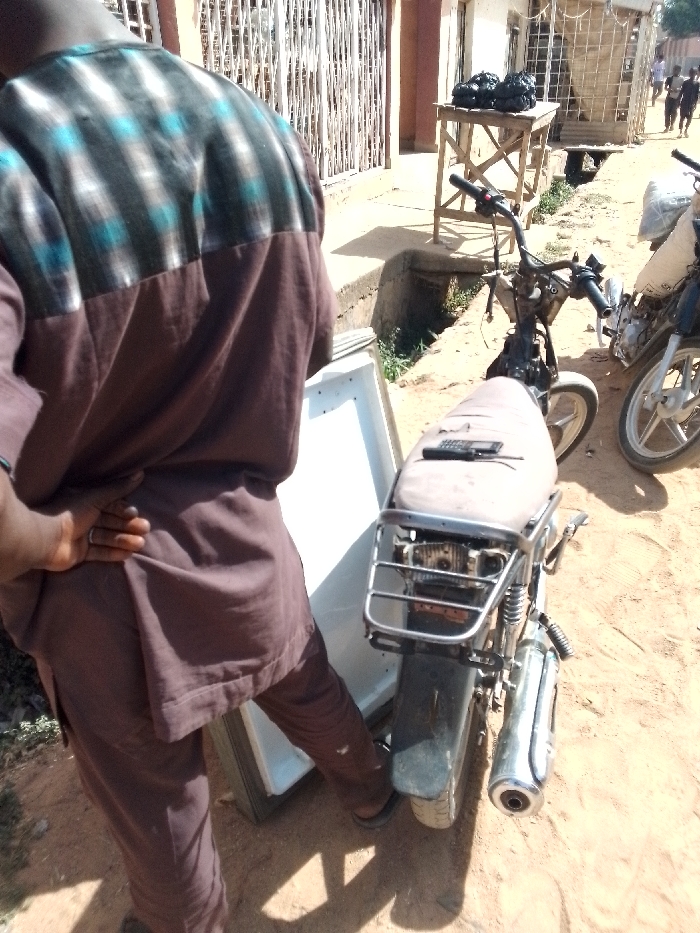 The image captures an outdoor scene, likely a street or pathway in a warm, dusty environment, during bright daylight. The ground is unpaved, consisting of dirt and sand.
In the foreground, a person is seen from the back, predominantly occupying the left side of the frame. They are wearing a traditional-style tunic or long-sleeved shirt and matching trousers, both in a solid brown or maroon color. The upper part of the tunic features a distinct pattern with horizontal and vertical stripes in dark brown/black, light brown, and light blue. The person's right hand is visible, resting on their lower back or hip, with a simple silver-colored ring on one of their fingers. Their posture suggests they are standing or possibly pushing/stabilizing a large, white object positioned directly in front of them. This object appears to be a refrigerator or freezer, lying on its side.
To the right of the person and the white object, a motorcycle or scooter is parked, seen from its rear and side. It appears to be an older model, possibly modified or partially disassembled, with visible engine components and a metal rack over the rear fender. A small, black feature phone is resting on its brown, worn seat. Another motorcycle is partially visible further to the right, showing parts of its front wheel and engine.
In the midground and background, the scene continues along a dusty path. A drainage ditch or culvert runs parallel to the path, containing some sparse green vegetation. On the far side of this ditch, a crude wooden table or stand is visible, topped with two black bags or sacks. Behind this, a building with barred windows can be seen. Further down the path, several indistinct figures of people are visible in the distance, walking or standing, indicating a populated area. The strong sunlight casts clear shadows, suggesting it is mid-day.