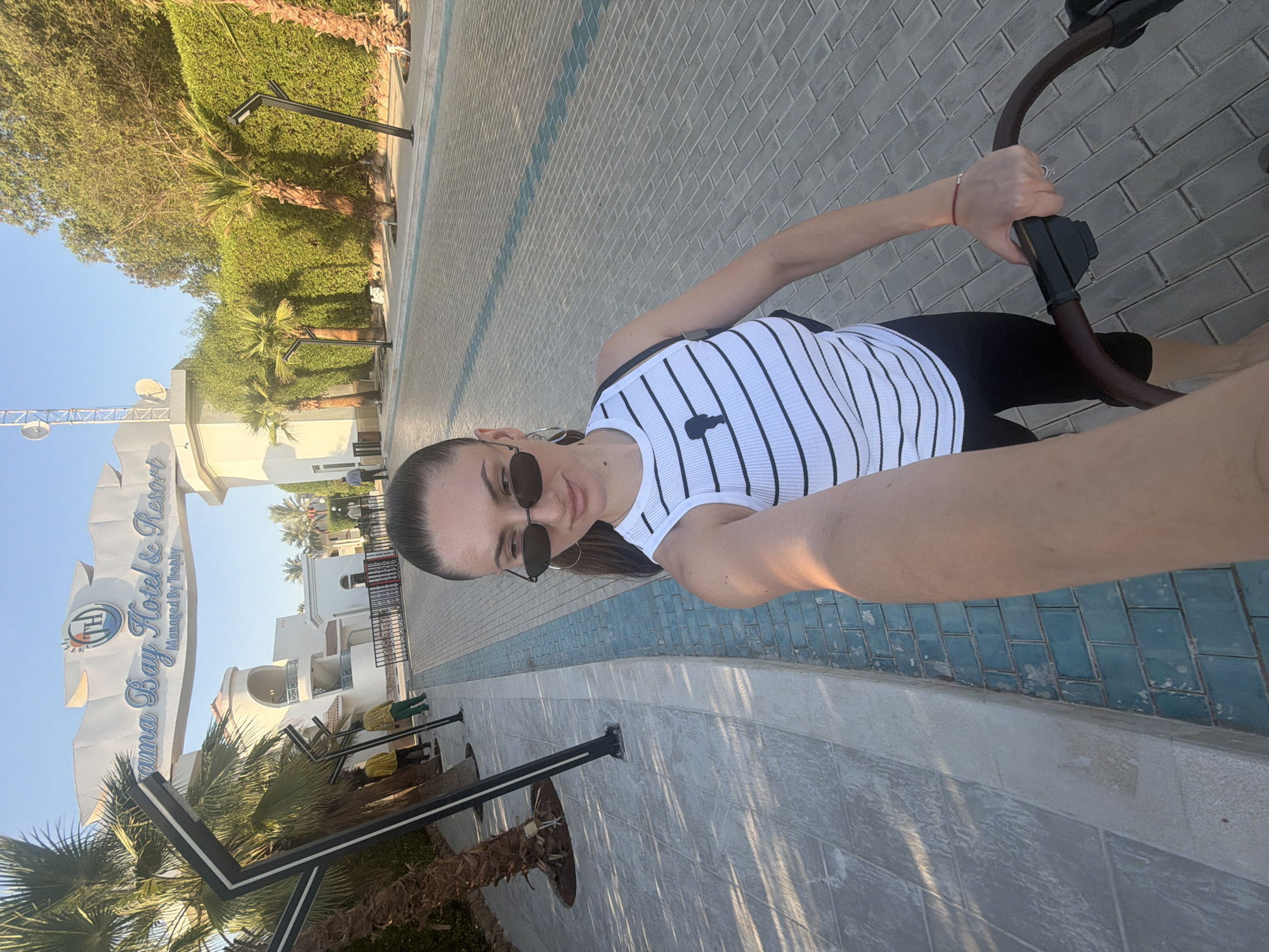 A woman with dark hair pulled back in a ponytail, wearing sunglasses, a white ribbed tank top with black horizontal stripes and a small black graphic, and black leggings, pushes a stroller. She is standing on a paved walkway in front of an archway that reads "Sharm Bay Hotel & Resort Managed By Trophy." The archway is white with blue lettering and a circular logo featuring a sun and the letters "TH." Behind the archway are buildings with white facades and balconies. Palm trees and lush green hedges line the walkway. The ground is paved with grey rectangular bricks, with a blue tiled border. The lighting suggests it is daytime, with clear skies and sunlight casting shadows. Two individuals in yellow shirts and one in green pants are visible in the background near the entrance.