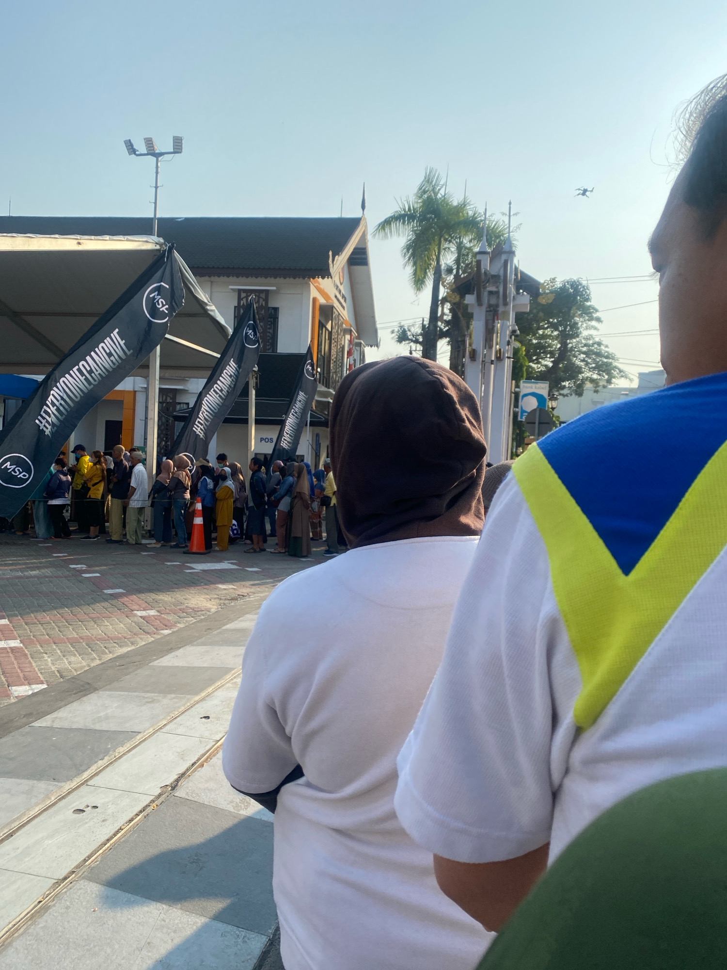 The image captures an outdoor scene with a clear sky, likely taken during the day in Banjarmasin, Indonesia. In the foreground, the backs of three individuals are visible. The person on the right wears a white shirt with a blue and yellow V-shaped design on the back. The person in the center wears a white shirt and a brown headscarf. A third person, mostly out of frame and to the left, also wears a white garment.
Behind these individuals, a line of people is formed, suggesting a queue. They are standing on a paved area with a textured, brick-like pattern. Several black flags with white text and circular logos reading "MSP" and "#EVERYONEGIVE" are planted along the queue. One orange traffic cone is visible near the people.
In the middle ground, a building with a dark roof and white walls is present. A sign with the word "POS" is visible on the building. Palm trees and other foliage are seen behind the building, along with a white structure with decorative elements. Overhead, a street light fixture is mounted on a pole. Further in the sky, a drone is captured in flight. The overall environment suggests a public space, possibly an event or a service point.