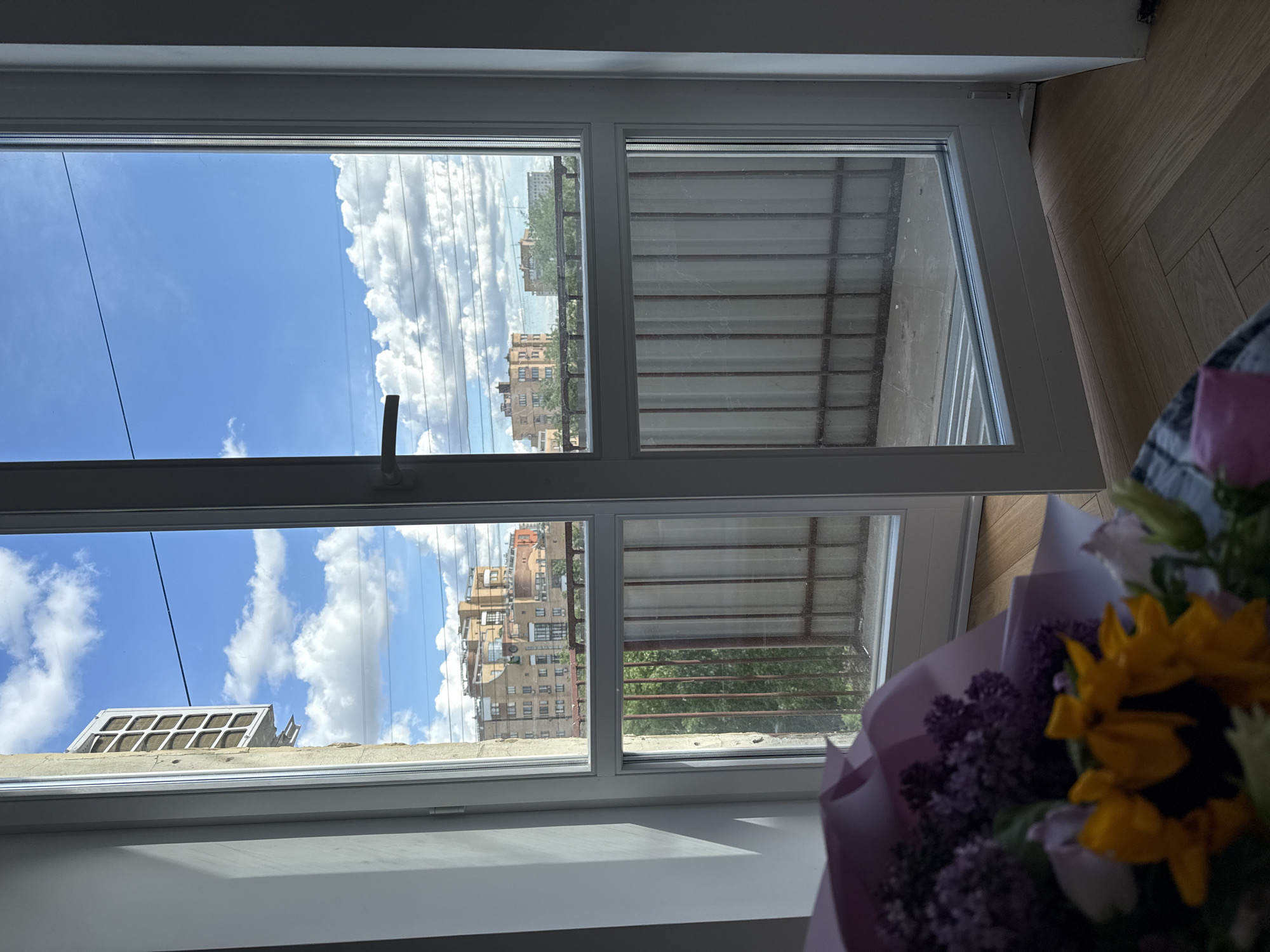 A view from inside a room, through an open white window, reveals a city scene under a bright blue sky with scattered clouds. On the left side of the window, the reflection of buildings and clouds can be seen. The window is partially open, showing a metal structure, possibly a balcony or similar feature. In the foreground, a bouquet of flowers, including sunflowers and purple flowers, is partially visible. The image captures a moment of everyday life, blending indoor and outdoor elements. The exact location in Moscow cannot be determined from the image.