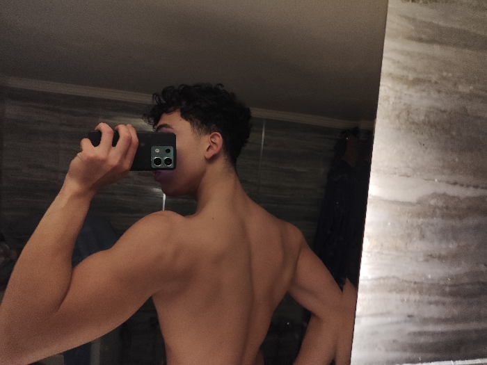 An indoor scene, identified as being in As-Salihiya Municipality, Syria, depicts a shirtless individual taking a mirror selfie. The person, appearing to be a young male with dark, curly hair and a fade haircut, is positioned with their back partially towards the viewer and their reflection visible in a large mirror that occupies the right side of the frame. The individual's left hand holds a black or dark-colored smartphone horizontally, with its prominent rear camera module (featuring two large lenses, a smaller third element, and a flash) visible, directed towards their reflection. Their right arm is flexed at the elbow, showcasing muscle definition in the bicep and tricep. The environment behind the person, as seen in the reflection, consists of dark, horizontally striped or tiled walls. A vertical segment of a lighter, possibly marble-patterned wall or partition is visible on the far right of the scene.