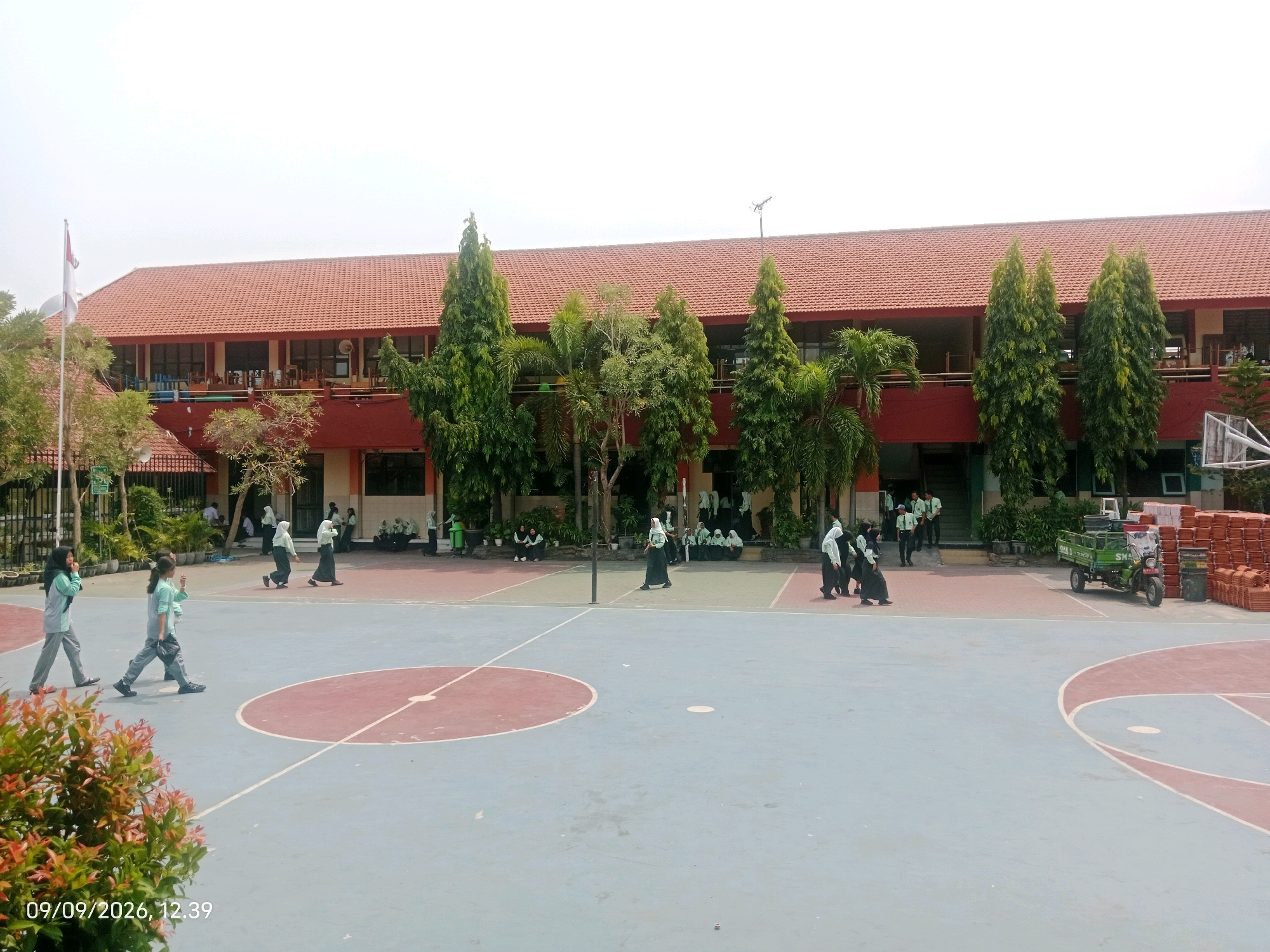 In an outdoor schoolyard, students in light green uniforms and dark headscarves are gathered on a basketball court. The court is marked with red and blue areas. Several students are walking across the court, some individually, and others in small groups. In the background, a two-story building with a red tiled roof and red walls stands. Trees are planted in front of the building. To the right, a green three-wheeled vehicle and stacks of terracotta roof tiles are visible. A basketball hoop is present on the far right. The sky is overcast. A timestamp reads "09/09/2026, 12:39".