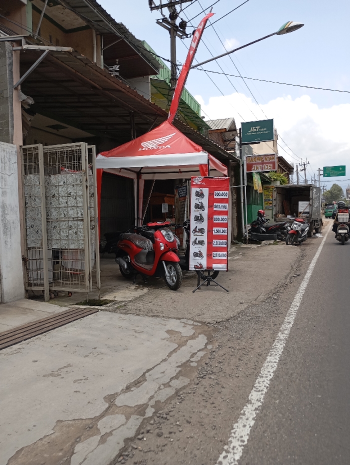 The scene is taken on a street corner in Katapang, Indonesia (latitude: -6.992, longitude: 107.556). A red and white canopy tent labeled "Honda" is set up on the sidewalk in front of a building. Inside the tent, a red motorcycle is parked. A sign next to the tent shows prices for motorcycles. The street is paved with asphalt and has a white line painted down the center. A few other motorcycles are parked along the street, and a truck is driving away in the background. There are several buildings along the street. The sky is partially overcast. The image was taken during the daytime.