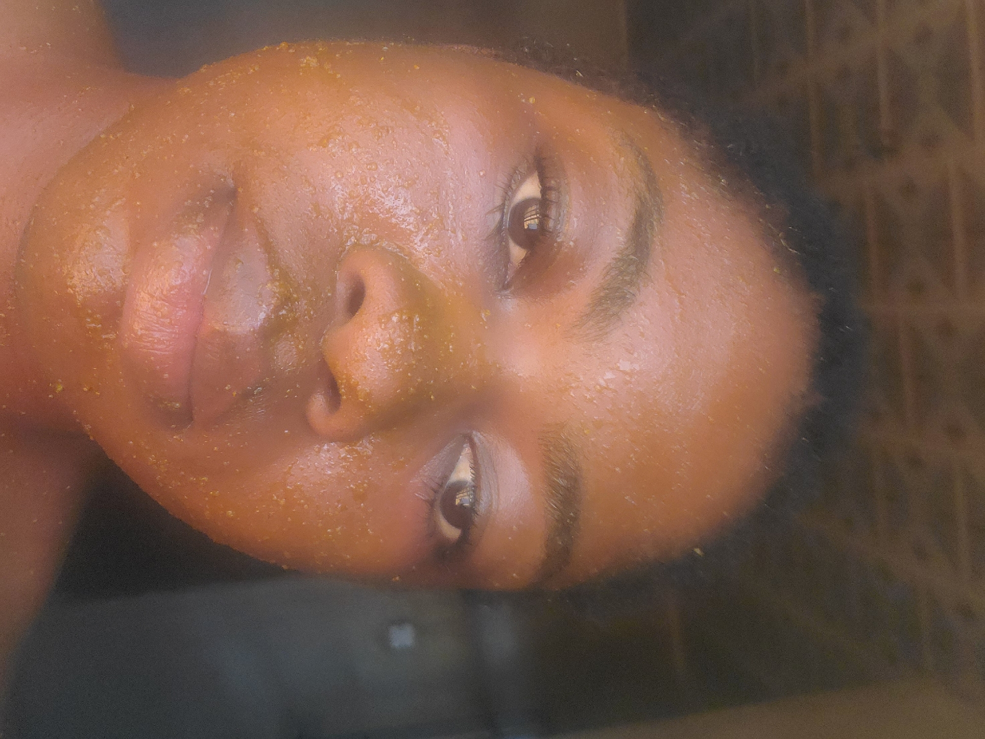 A close-up portrait of a dark-skinned individual, likely a young adult female, is depicted in Kakau, Nigeria. The subject faces the camera directly, maintaining eye contact, with a neutral expression. Their face is entirely coated in a granular, yellowish-orange substance, consistent with a facial mask or scrub, covering the forehead, cheeks, nose, and chin. Small specks of the substance are visible along the hairline and upper chest. Both eyes are open, and reflections of an unidentified light source are present in the pupils. The hair, dark and short, is visible at the top of the head. The background is dark, out-of-focus, and appears to be an interior wall or doorway with subtle, barely discernible horizontal textures. The lighting is frontal and bright, illuminating the subject against the subdued background.