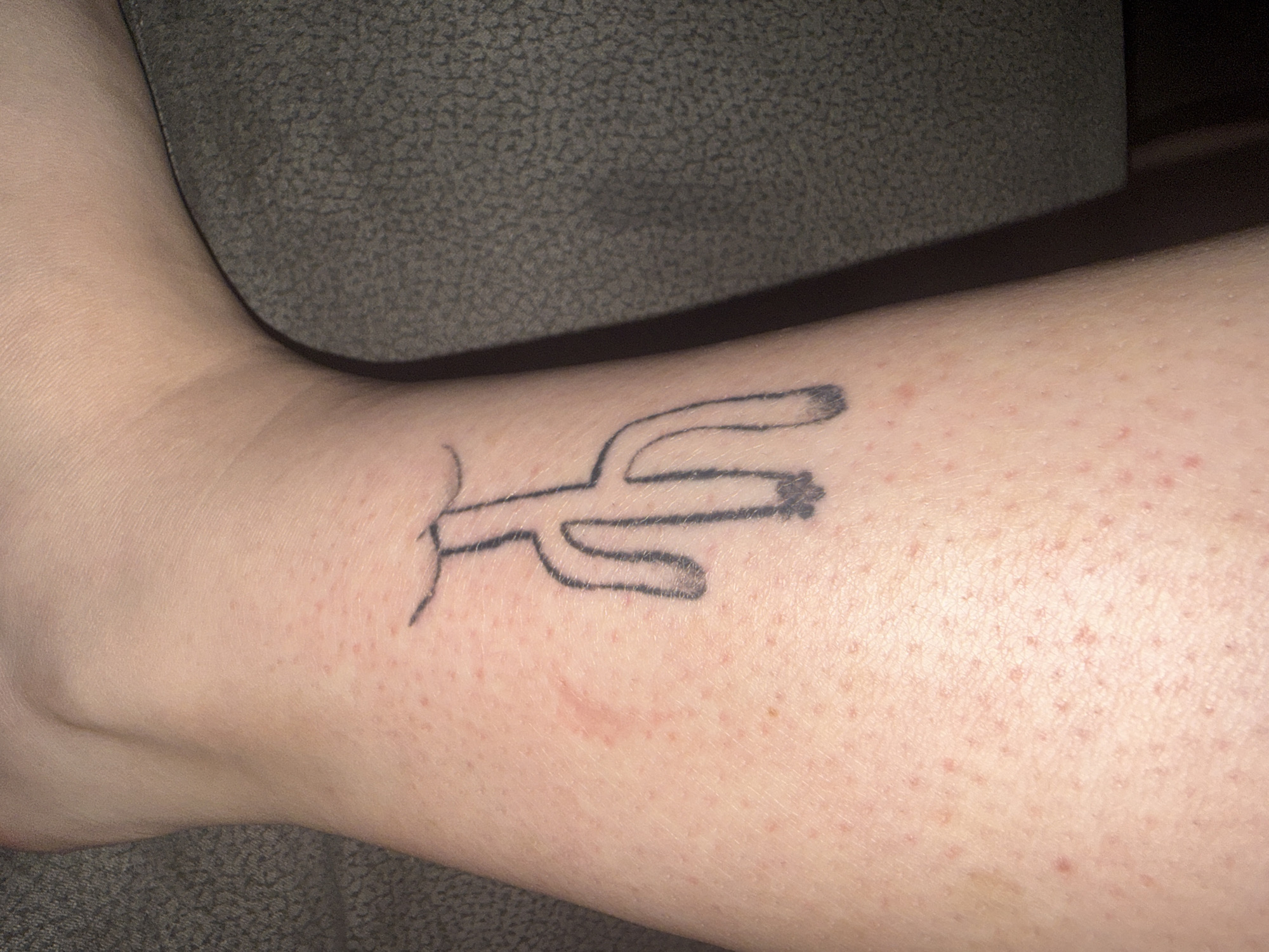 A black line tattoo of a simplified, abstract figure resembling a cactus or a stick figure with large arms and legs is visible on a person's leg. The tattoo is located on the lower part of the leg, near the ankle, on skin that appears fair and has some small red blemishes. The background is a dark, textured surface, possibly upholstery.