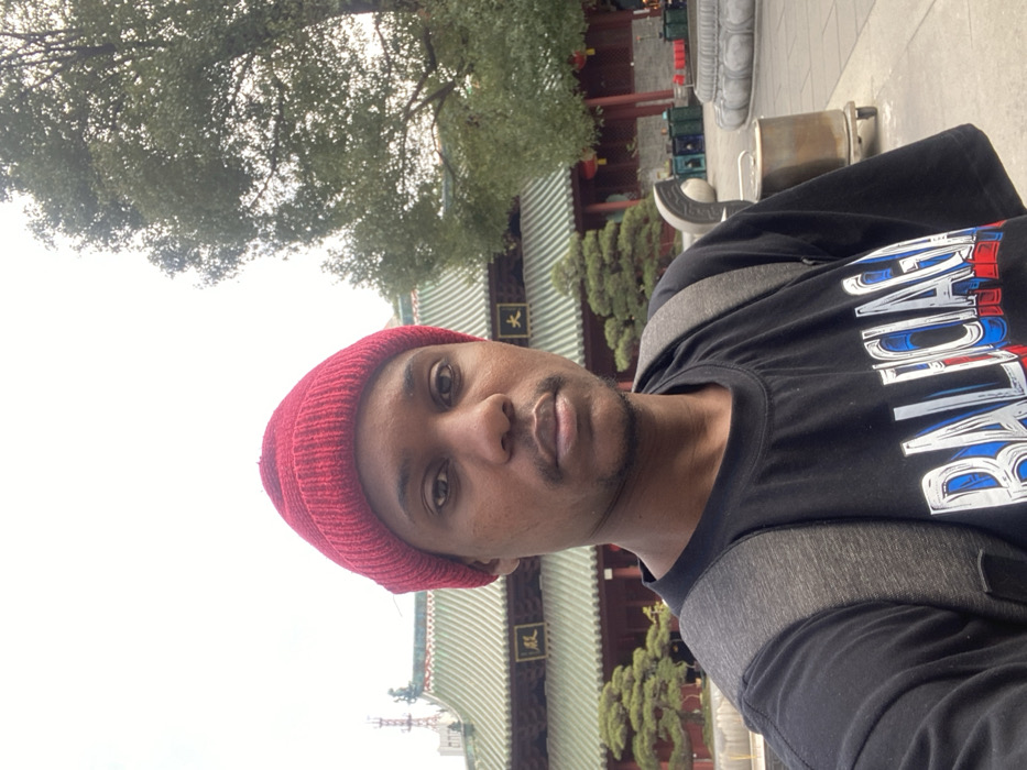 A selfie captures a young Black man outdoors in what appears to be a traditional Chinese temple complex in Guangzhou, China. He looks directly at the camera with a neutral expression. He wears a red knit beanie and a black t-shirt with "BALENCIAGA" written across the front in a stylized font with blue and red outlines. Grey backpack straps are visible on his shoulders.
The background features striking traditional Chinese architecture, including a large building with an intricate green tiled roof and red pillars. On the building's facade, vertical Chinese characters are visible, likely reading "天王殿" (Hall of Heavenly Kings), a common structure in Buddhist temples. Surrounding the architecture are lush green trees, including a dense canopy overhead and manicured bushes. Stone railings and what appears to be a traditional incense burner or offering pot are also present near the building. The lighting is diffused, suggesting it is daytime under an overcast sky.