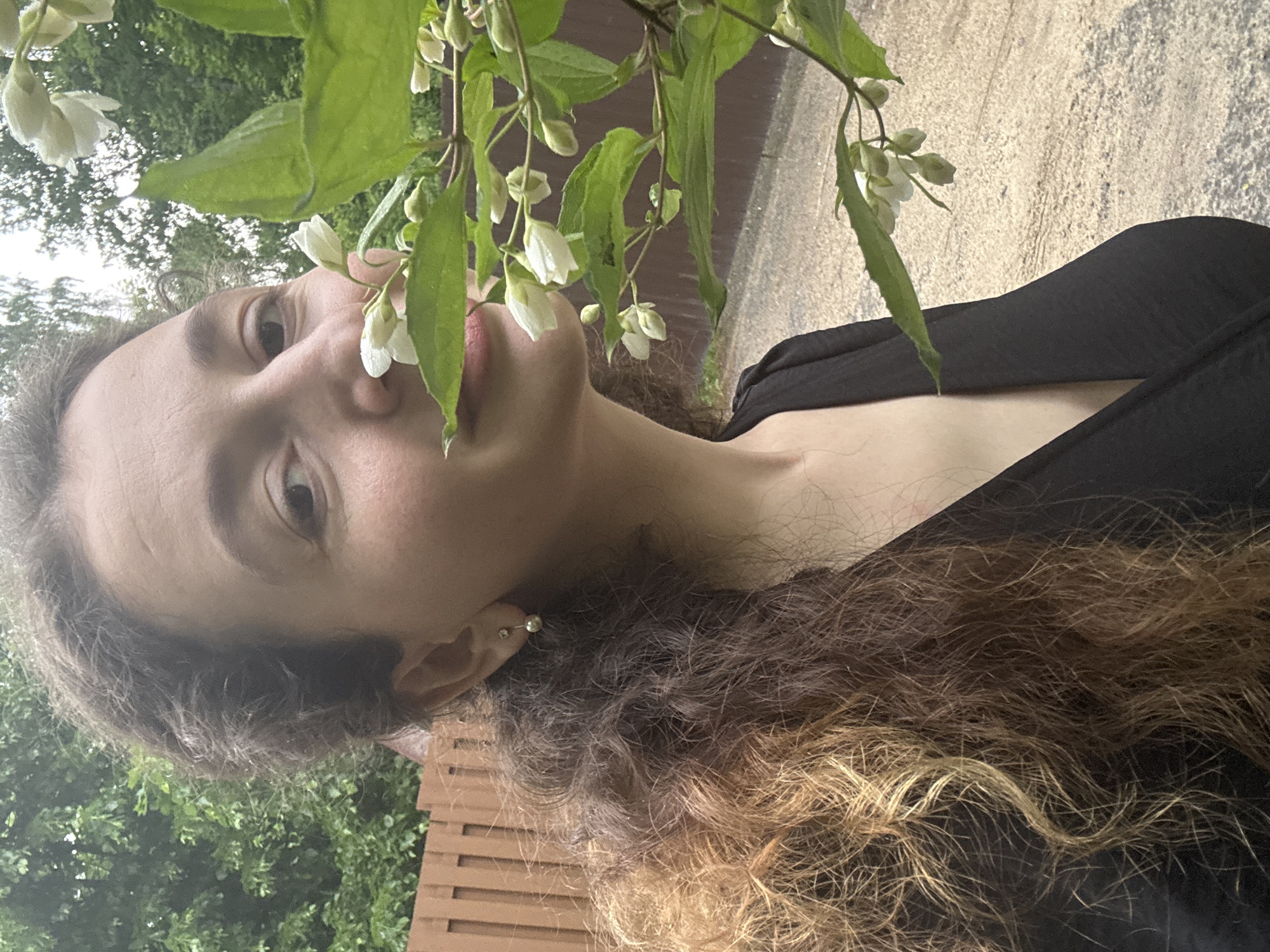 A medium shot captures a woman from the chest up, positioned in a shallow focus outdoor setting. She is facing the camera, with her right side slightly closer. Her dark, curly hair, streaked with lighter brown tones, cascades down her left shoulder. A subtle pearl and ball earring is visible on her right ear. She wears a black V-neck top. Her gaze is directed towards the viewer, and her lips are slightly parted as she inhales the scent of a white-flowered plant in the foreground, which partially obscures her face. The plant's green leaves and white blossoms are in sharp focus. Behind her, a wooden fence runs horizontally across the background, with a blur of green foliage and trees further beyond. The ground appears to be a light-colored gravel or dirt path. The overall lighting suggests an overcast day.