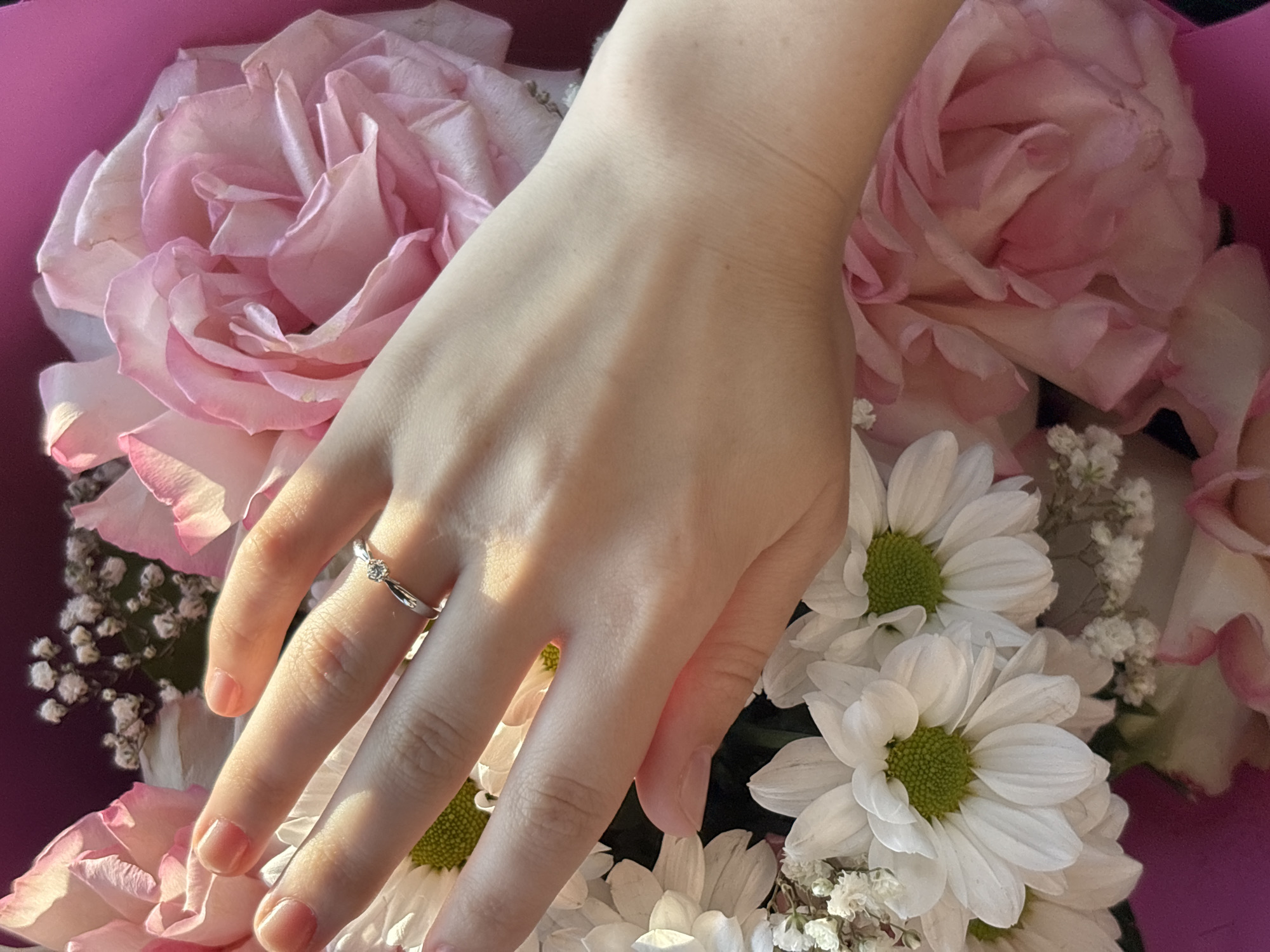 A close-up shot shows a woman's hand, adorned with a delicate ring featuring a small diamond, resting on a bouquet of pink roses and white daisies. The hand is positioned at an angle, with the fingers slightly curled. The sunlight casts shadows and highlights on the skin, emphasizing its texture. The bouquet is lush and vibrant, with soft pink petals of roses contrasting with the crisp white of the daisies. The background is a blur of pink and dark purple, suggesting the flowers are arranged in a bouquet. The setting does not provide any information about its location in Saint Petersburg, Russia.