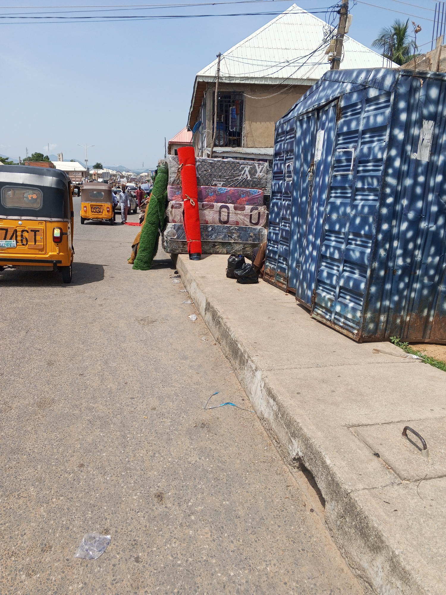 This is a street scene in Jalingo, Nigeria. In the foreground, a gray concrete curb runs along the right side of the asphalt road. On the road, several objects are visible. A yellow and black auto rickshaw, identified by the license plate "746T", is parked on the left. Further down the street, another auto rickshaw, with license plate "093L", is also visible. A stack of mattresses, covered in plastic wrapping with various brand names like "Louis Vuitton" and "O", is piled on the side of the road. A rolled-up red carpet and a roll of green artificial turf are placed next to the mattresses. Two black plastic bags are situated near the base of the mattresses. In the background, buildings line the street, and a blue shipping container with white speckles is positioned on the right side of the image. People are seen walking on the street and near the vehicles. The sky is clear and blue, suggesting a sunny day.