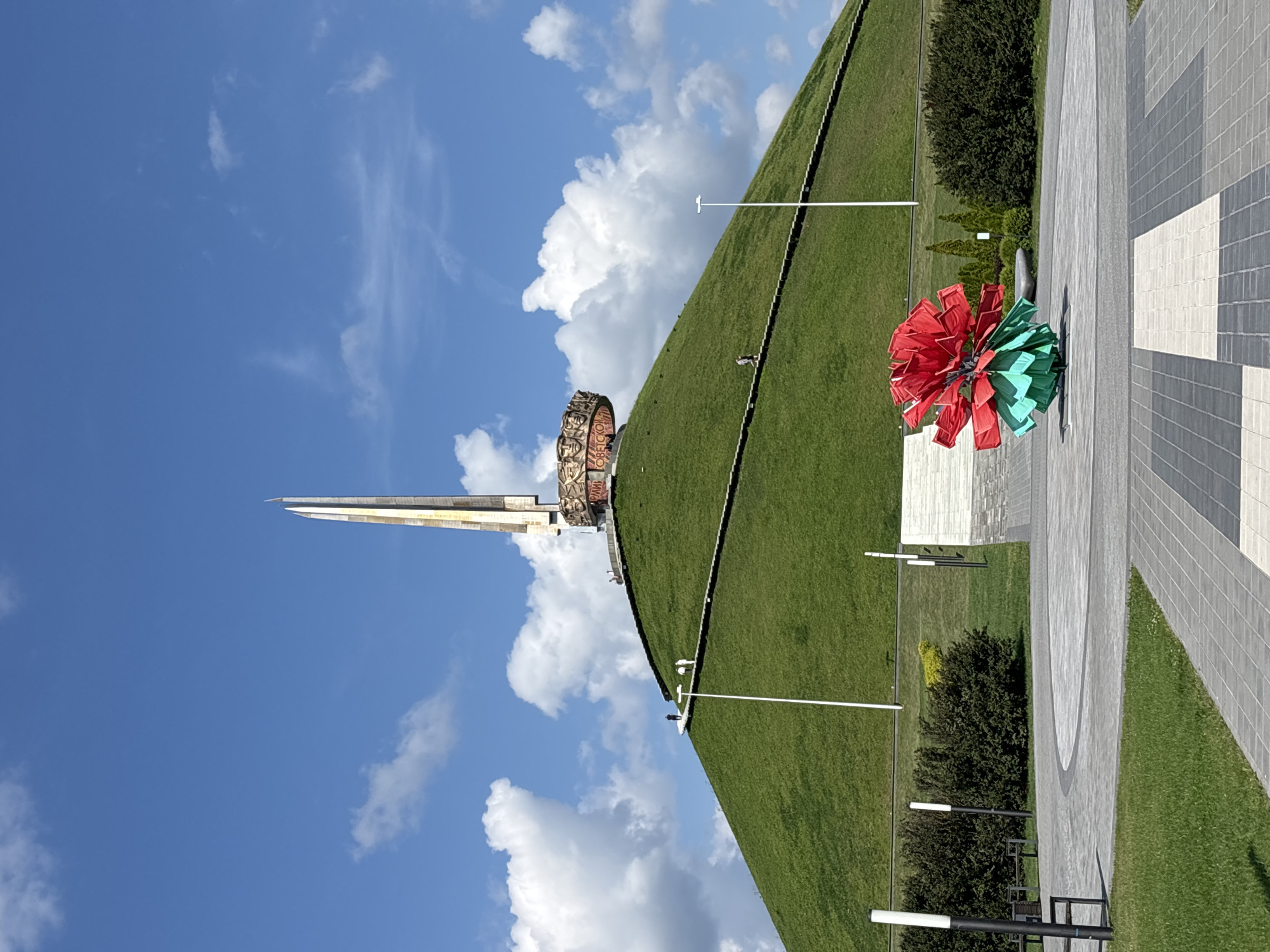 The image depicts the Mound of Glory, a prominent war memorial located in the vicinity of Ozyaritska-Slabadski rural council, Belarus. The memorial is situated atop a large, conical green hill, with a tall obelisk-like structure rising from its summit. The obelisk is adorned with intricate carvings and appears to be made of a metallic material. A circular frieze, also featuring relief sculptures, encircles the base of the obelisk. Stone stairs ascend the side of the hill, leading to the monument. Several individuals are visible on the stairs and near the base of the monument, suggesting it is a site of public interest. In the foreground, a colorful abstract sculpture resembling a large red and green flower is displayed on a paved plaza. The plaza is surrounded by manicured green lawns and dark green bushes. The sky above is a vibrant blue with scattered white clouds, indicating fair weather. Several lampposts are also present in the plaza area.