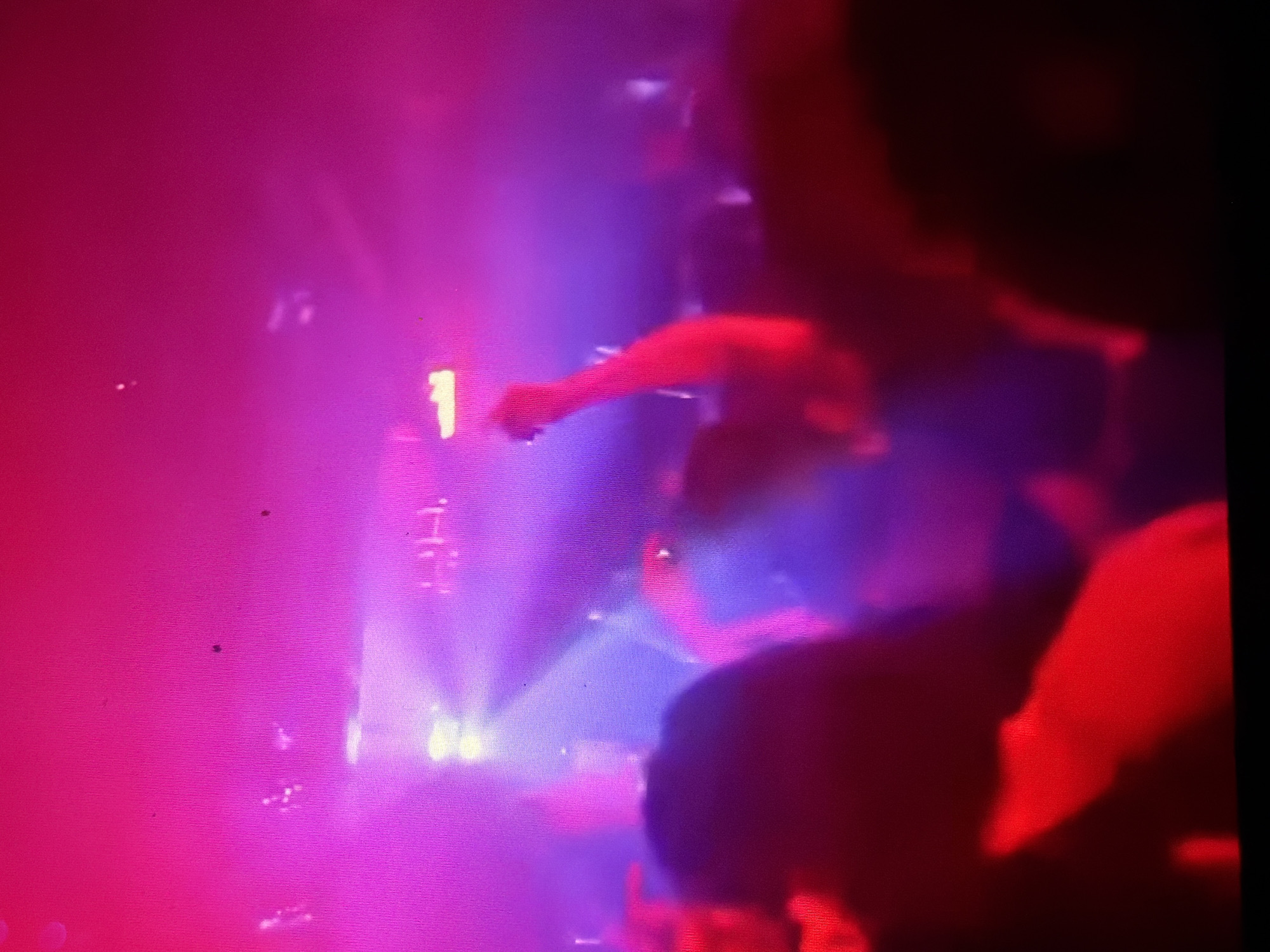 The image shows a dimly lit indoor scene with a crowd of people dancing. The lighting is predominantly pink and purple, with some yellow and red accents. Several individuals are visible, their features blurred by the low light and motion. Some people are raising their arms, suggesting they are actively participating in the event. In the background, there are visible lights that appear to be part of the venue's lighting system. The atmosphere suggests a nightclub or a similar entertainment venue. There is no identifiable information within the image to suggest a specific location in Oslo, Norway.