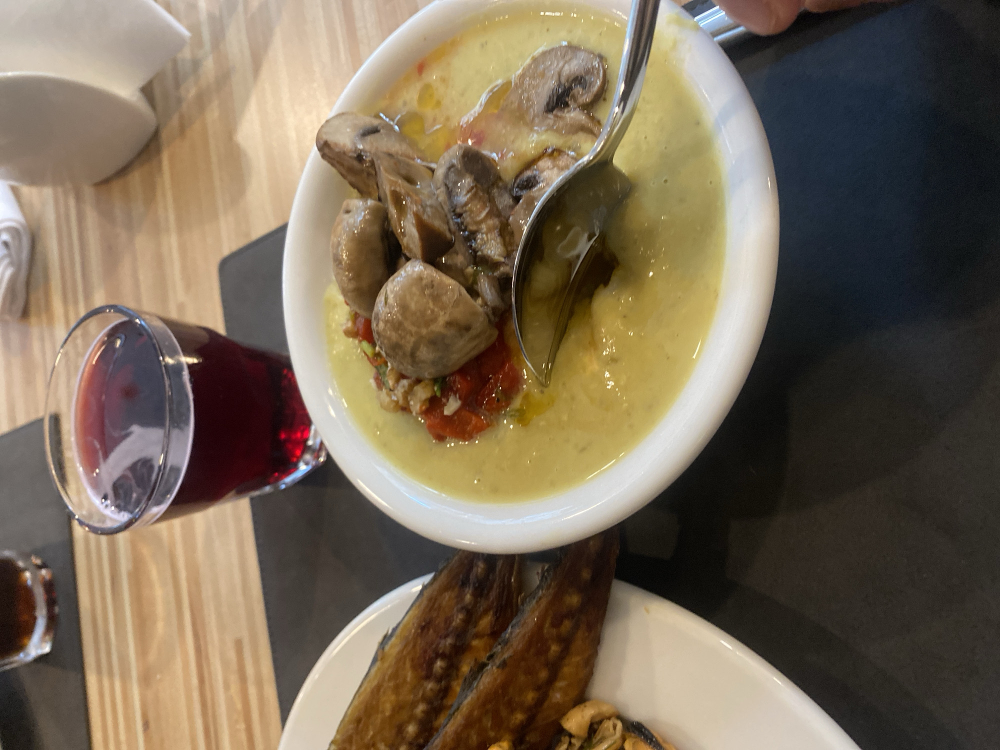 A close-up shot shows a white bowl filled with a creamy, light green soup topped with sliced mushrooms and red diced ingredients. A silver spoon rests in the soup, appearing to be in motion. To the left of the bowl, on a white plate, are two grilled fish, likely mackerel, arranged side-by-side. The scene is set on a wooden table with a black placemat. In the background, a glass filled with a dark red liquid, possibly wine or berry juice, sits on a dark coaster. Another out-of-focus glass of dark liquid is visible further in the background. A white paper napkin dispenser can be seen in the upper right corner. The lighting is ambient, suggesting an indoor setting. No people are clearly visible, though a hand is partially visible on the right edge of the frame. The image appears to be taken at a restaurant in Aksu, Turkey, given the context.