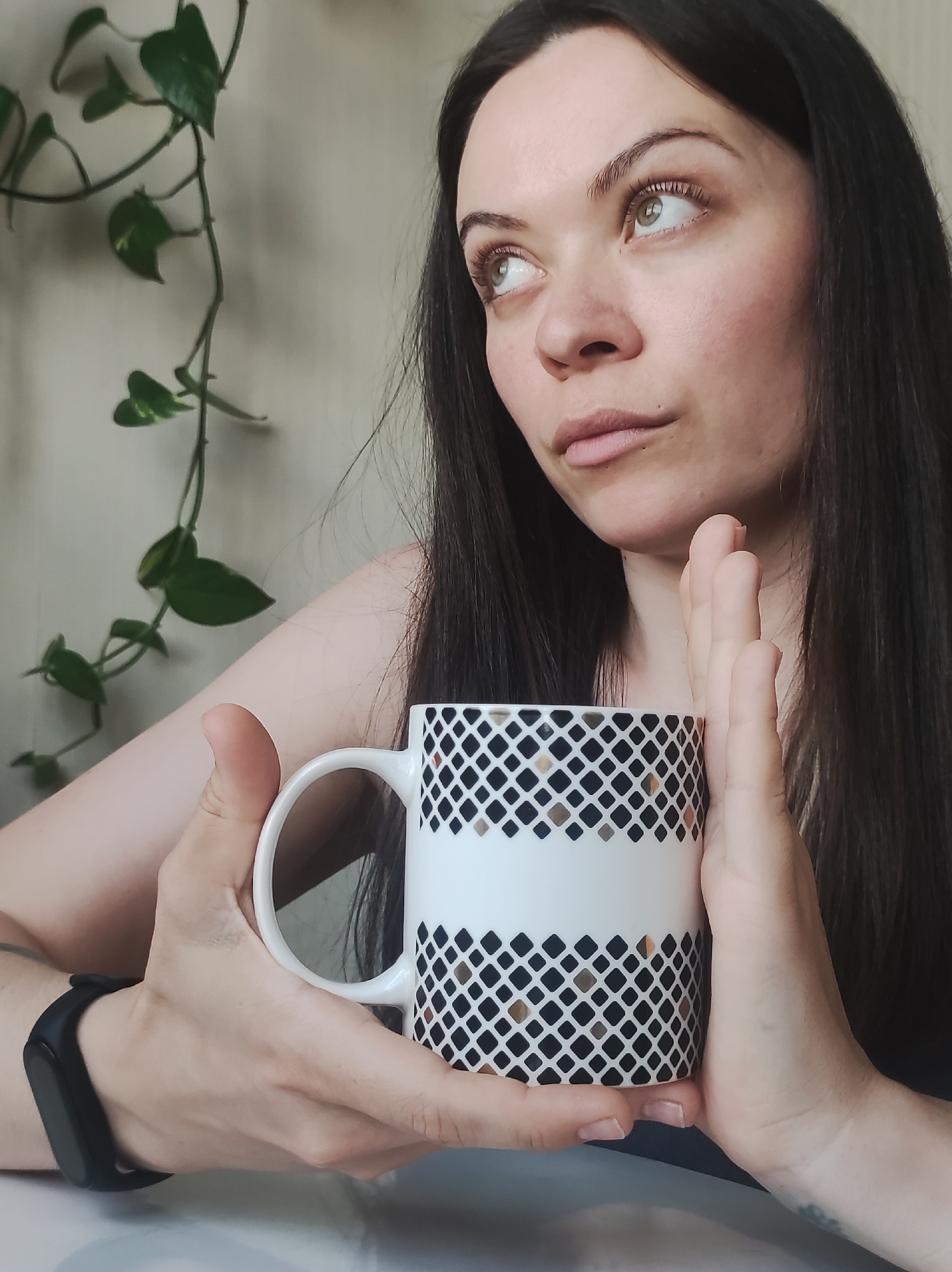 The image captures a close-up, chest-up view of a woman holding a mug with both hands. The woman has long, dark brown hair and light-colored eyes, possibly green or hazel, which are cast upwards and to the right, giving her a thoughtful or contemplative expression. She has fair skin and appears to be wearing a dark top. A black fitness tracker or smartwatch is visible on her left wrist.
She is holding a white mug adorned with a repeating geometric pattern of black squares, interspersed with some gold or bronze squares, forming horizontal bands. Her hands gently cradle the mug.
In the blurred background to the left, a green vine plant with heart-shaped leaves, possibly a Pothos, hangs against a plain, light-colored wall. The lighting appears soft and natural, suggesting it is daytime indoors.
The scene conveys a calm and introspective moment. There is no visible text in the image. The setting is an indoor space, located in Chaikovsky, Russia.