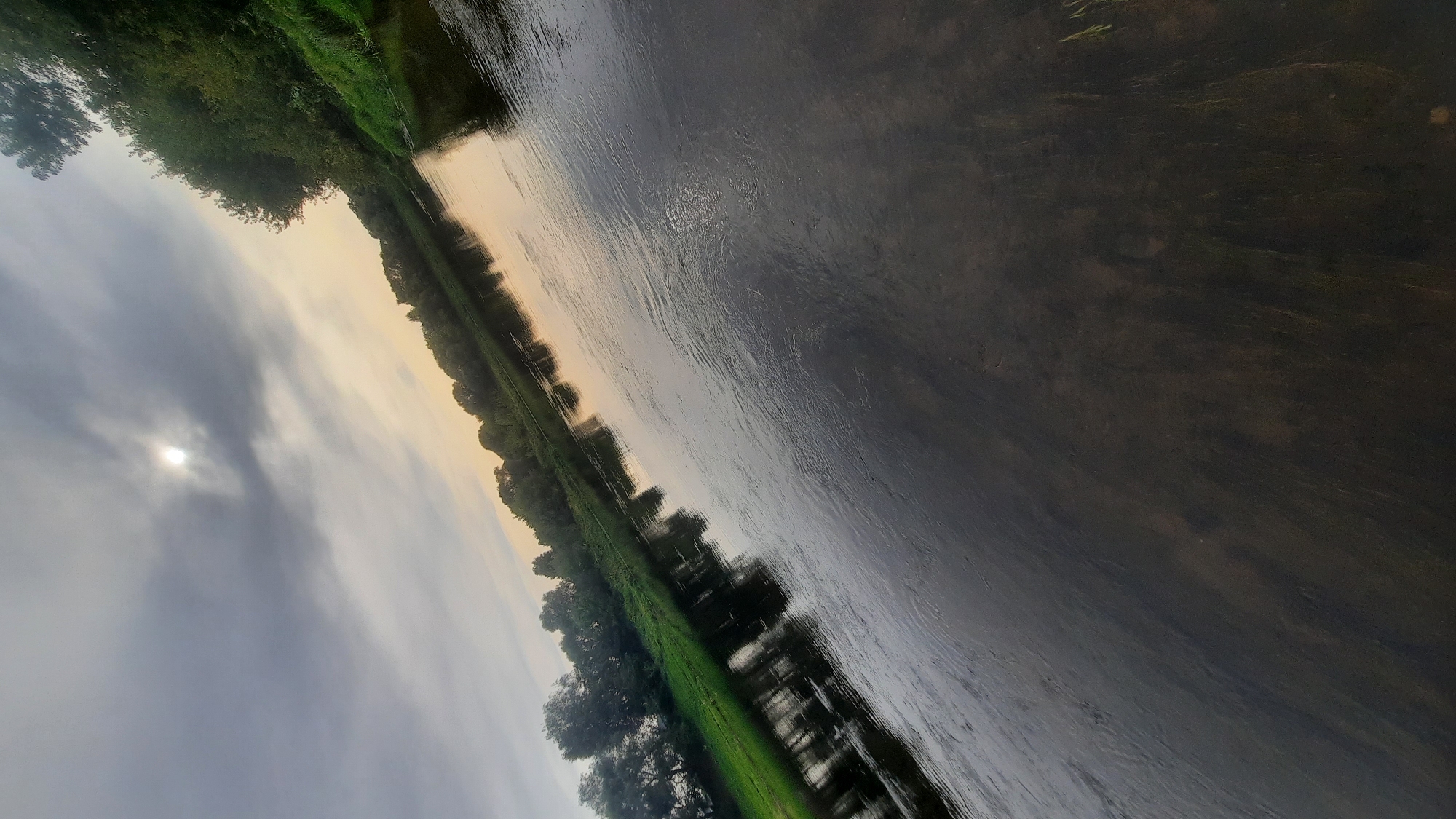 The image captures a natural landscape scene, likely a riverbank, with the sun partially obscured by clouds in the sky. The water occupies a significant portion of the frame, reflecting the surrounding environment. On the left side, a grassy bank is visible, dotted with trees and their reflections in the water. The right side of the image features a denser line of trees, also reflected. The overall impression is one of a serene, possibly overcast day. The location in the city of Шклоў, Belarus, suggests this is a natural area within or near the city. No people or specific actions are depicted.