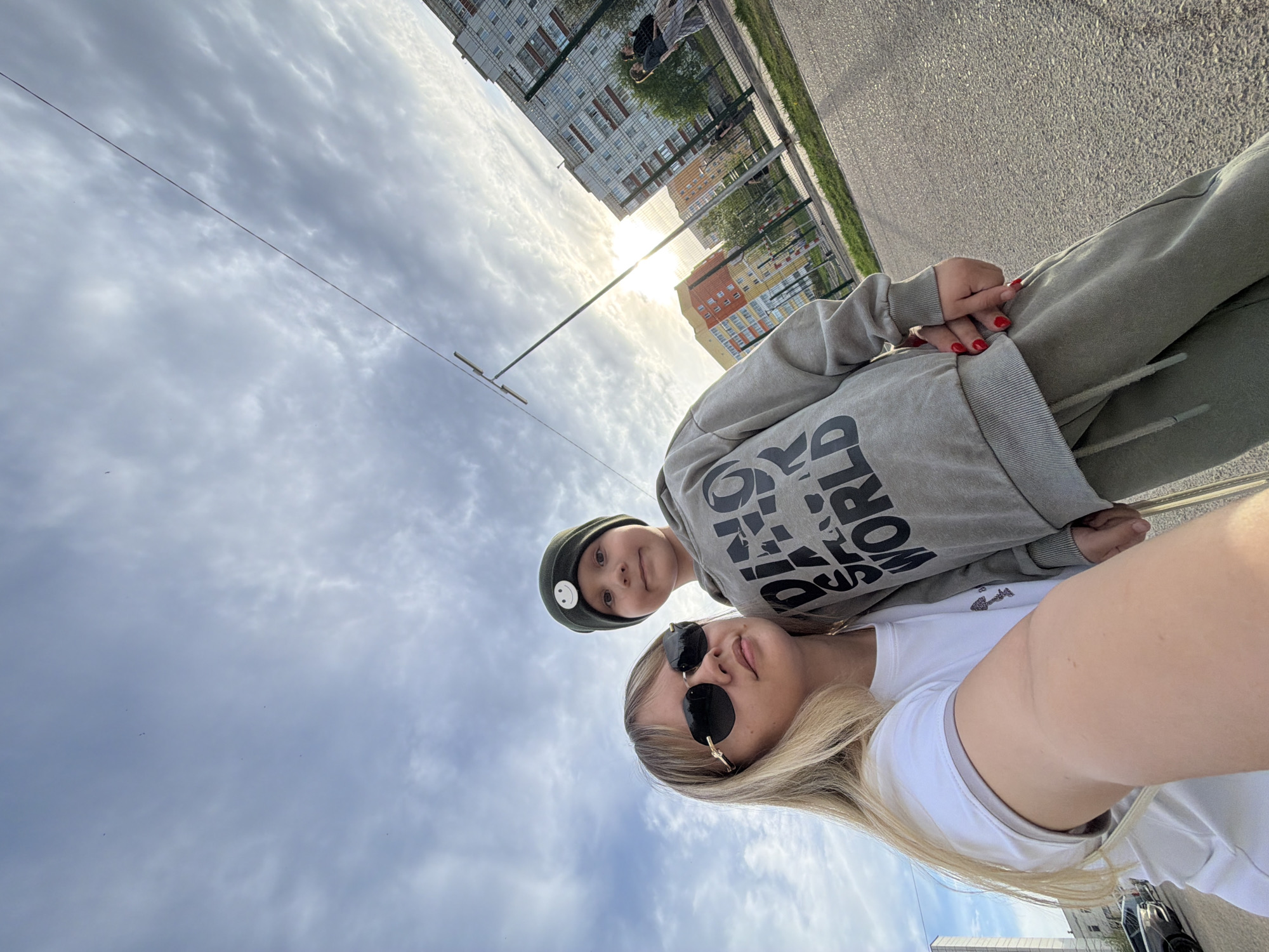 A woman and a child are posing for a selfie outdoors. The woman has blonde hair and is wearing sunglasses and a white shirt. The child is wearing a dark hat with a smiley face on it and a grey sweatshirt. They are standing on a paved area. Behind them are buildings, indicating an urban setting. The cloudy sky is visible in the background. Based on the appearance of the buildings and other environmental factors, it is likely that the location is Chaikovsky, Russia, as specified.